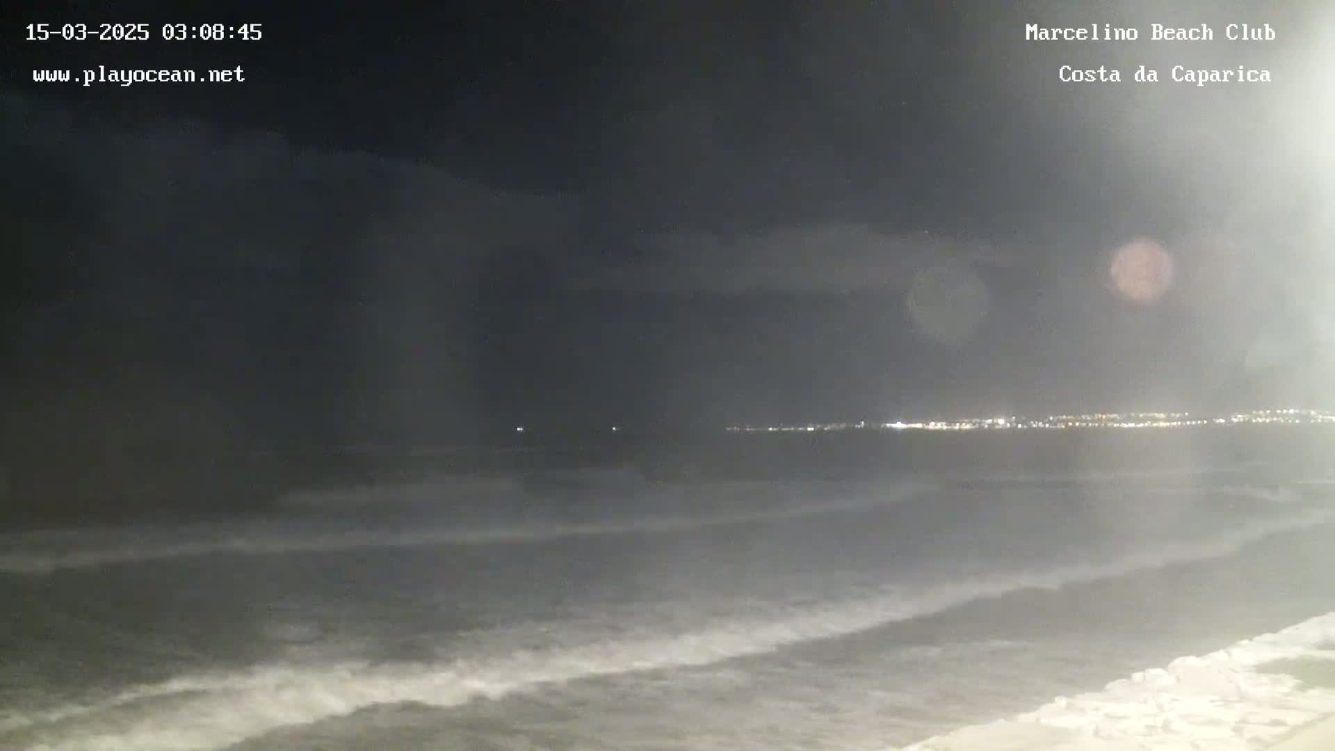 A dark nighttime view shows gently rolling waves on the ocean, with a distant city skyline faintly illuminated.