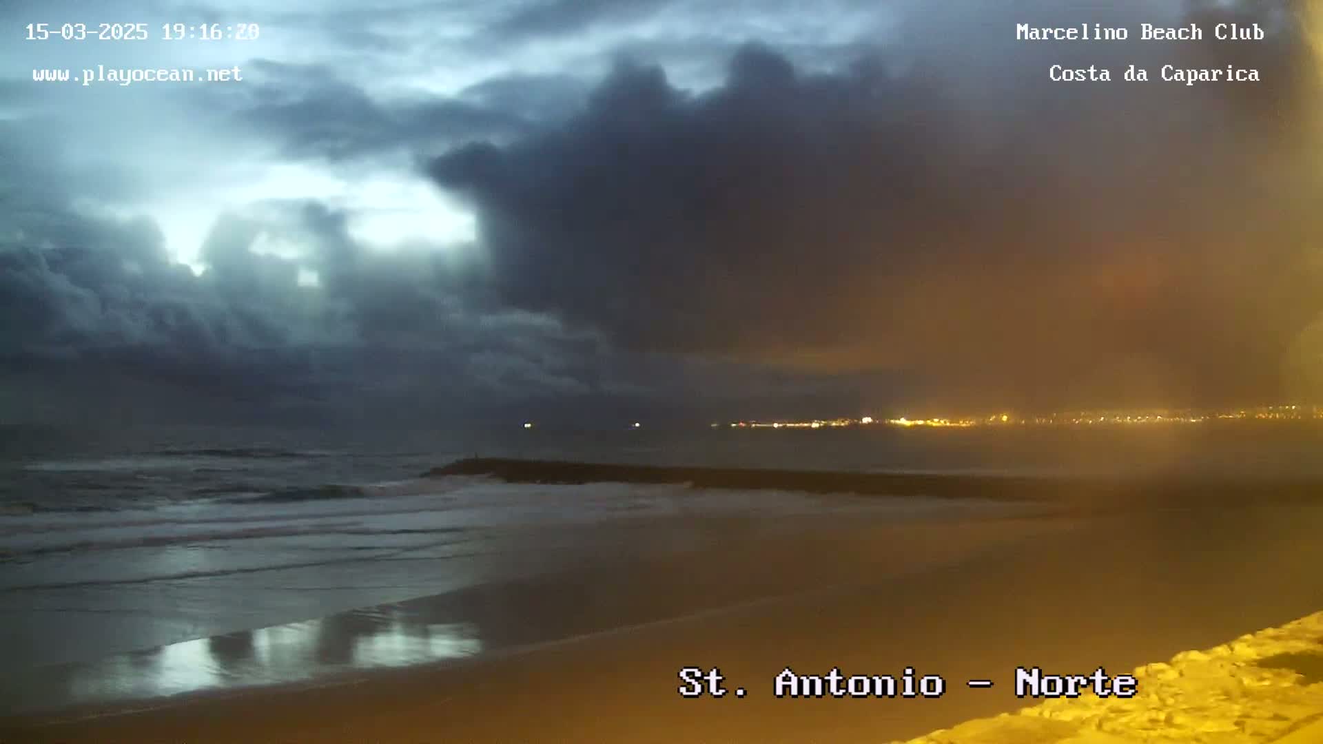 A dark, cloudy evening scene shows a beach with waves gently lapping the shore, under a sky filled with heavy, gray clouds.