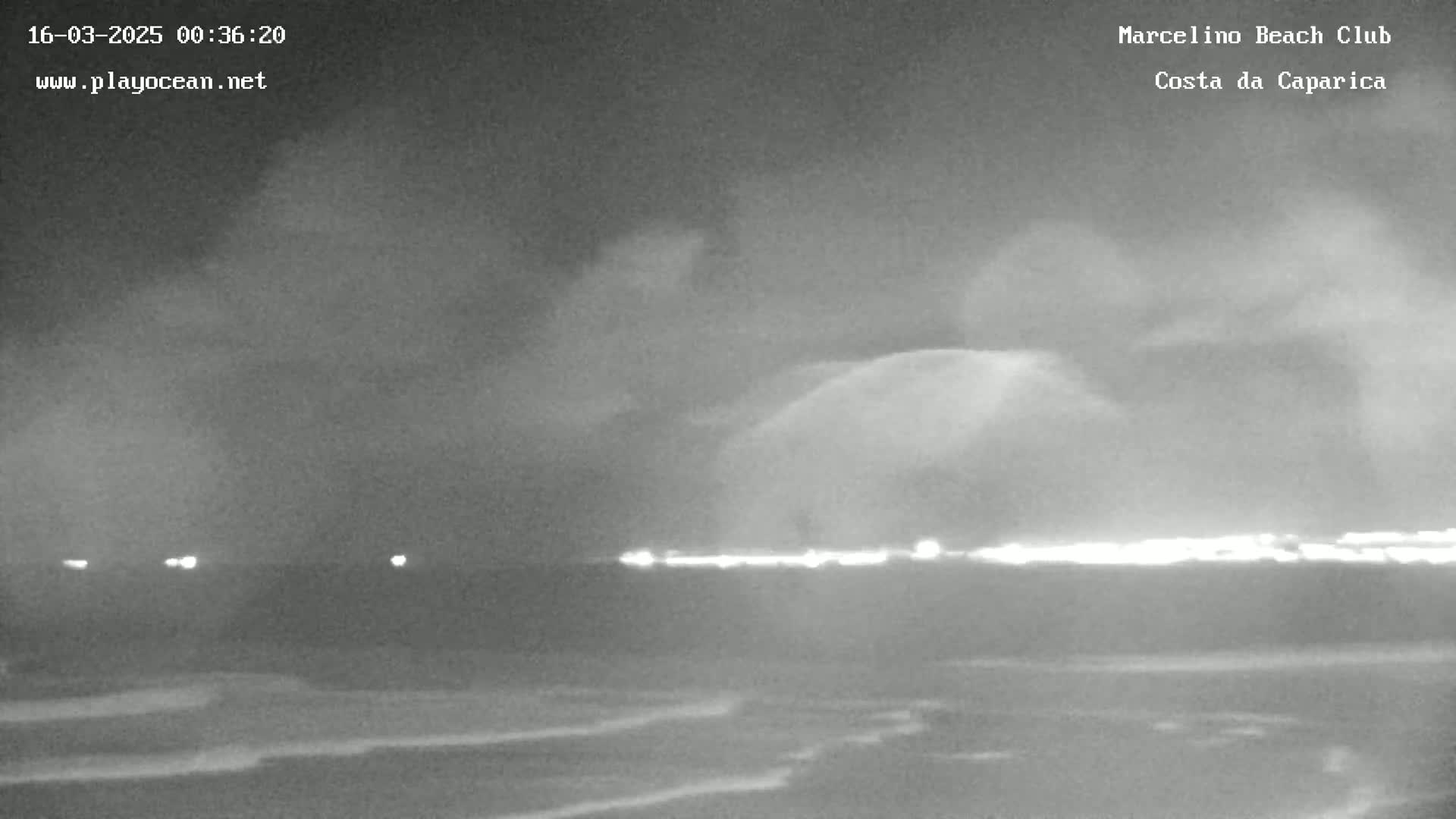 A nighttime, overcast view of a beach with a dark ocean and a faint line of lights on the horizon.