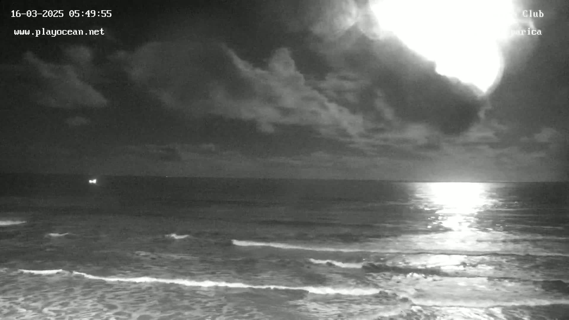 A nighttime ocean scene shows a bright light on the horizon under a partly cloudy sky with gentle waves.