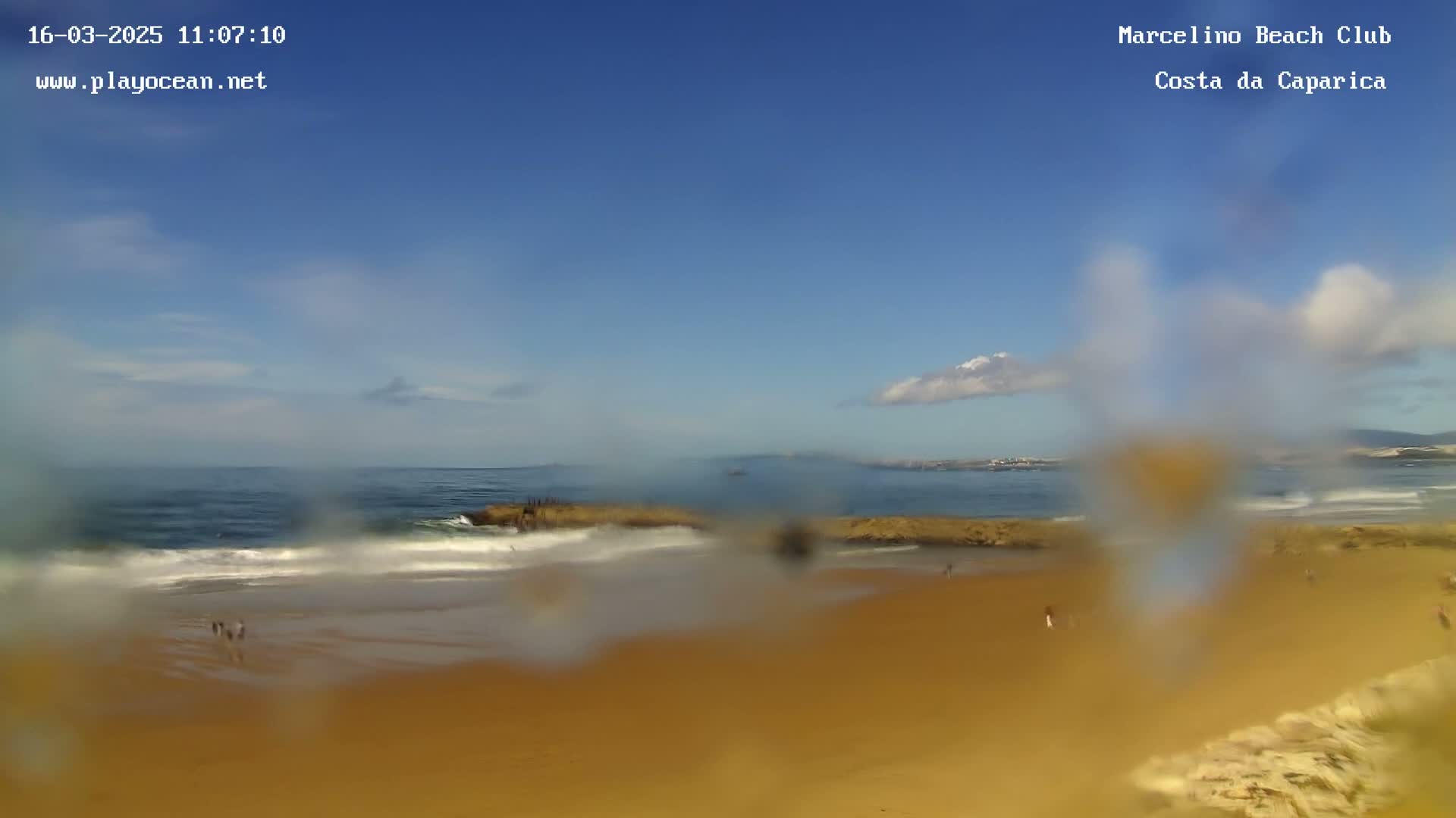 A mostly sunny beach scene with small waves rolling onto a sandy shore is viewed through a rain- or water-covered surface.