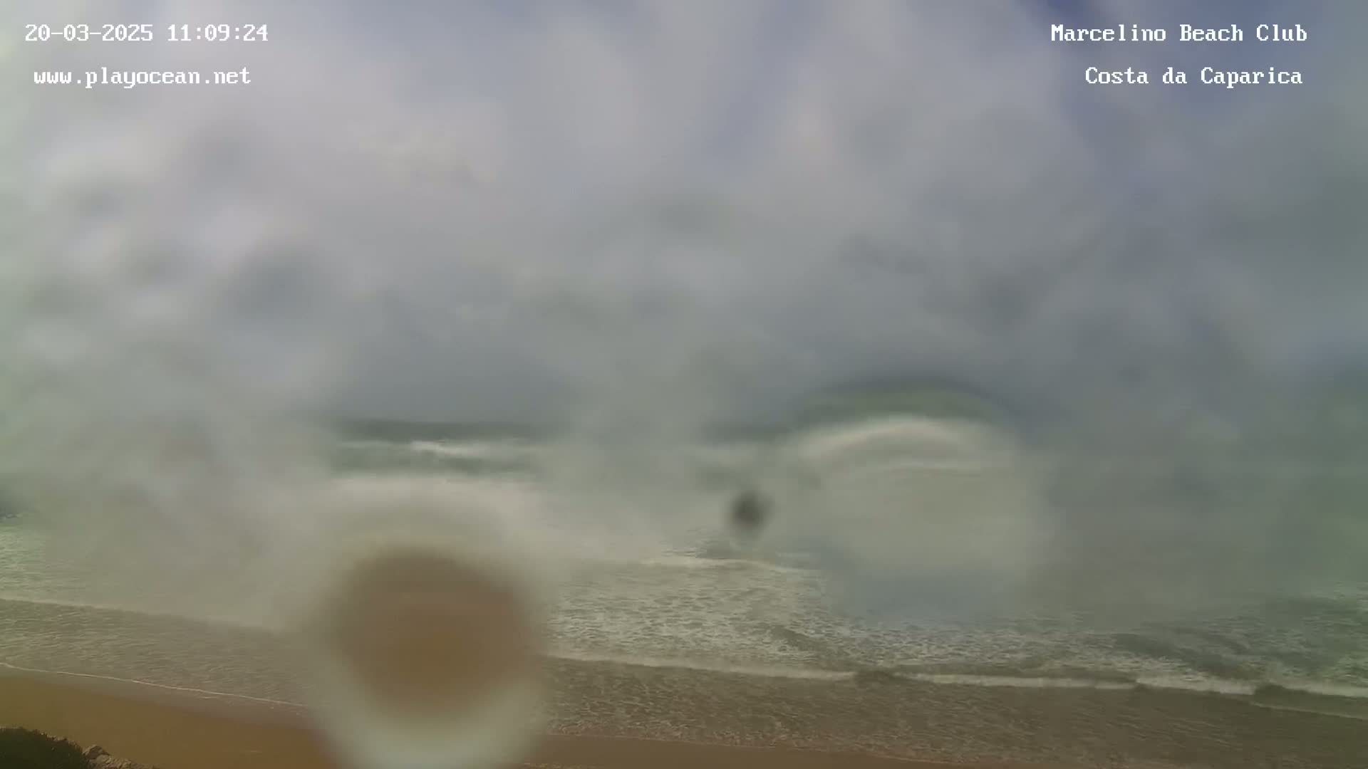 A hazy, overcast sky hangs over a beach with moderately sized waves rolling onto the shore.