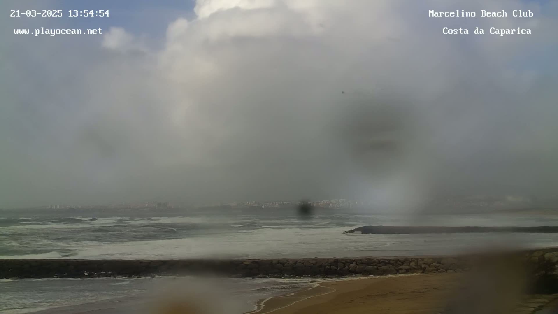A mostly cloudy and overcast day shows a beach with choppy waves and a distant city under a hazy sky.