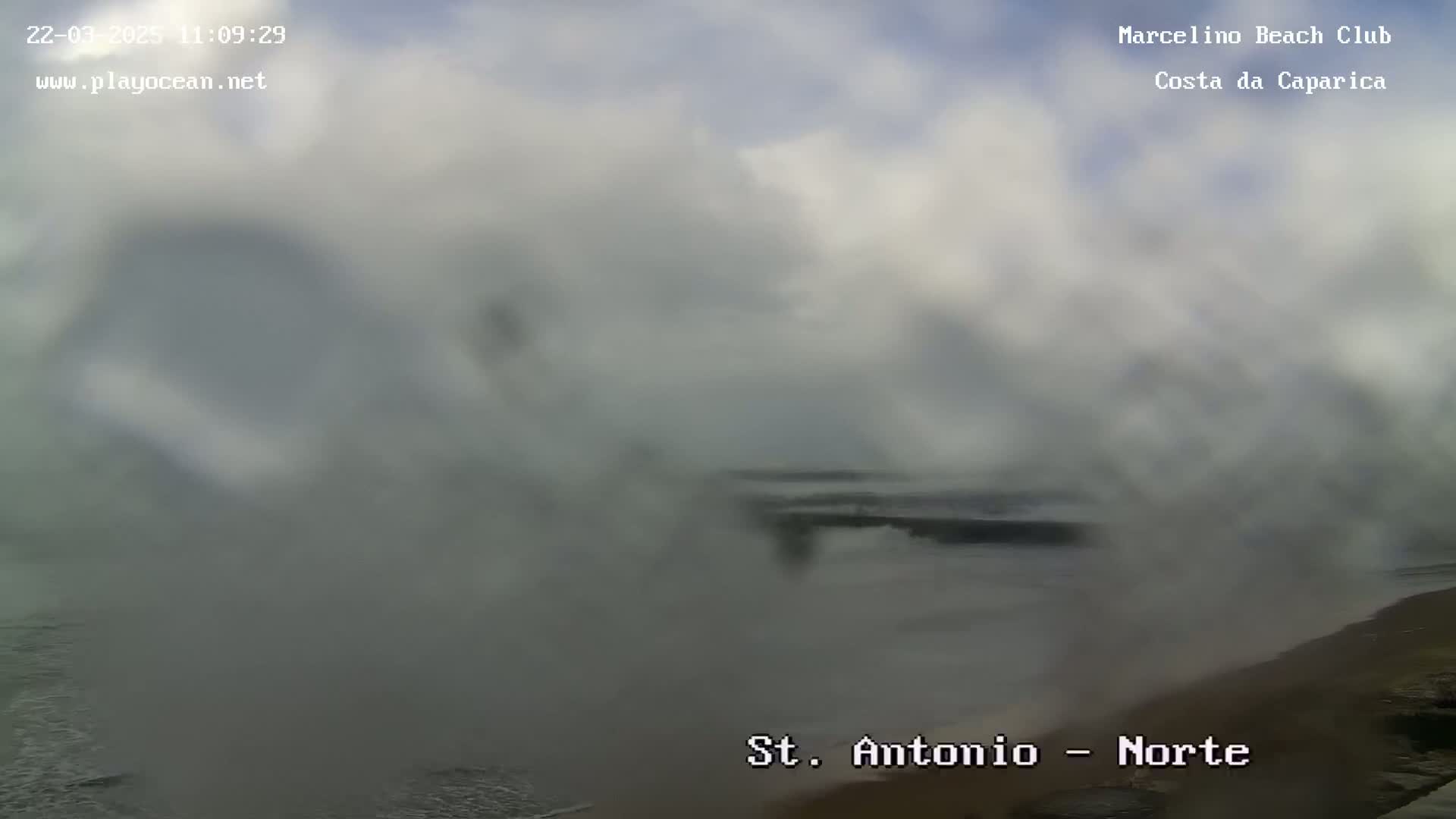 A hazy, overcast day at a beach shows a partially visible structure near the shore with waves crashing in the foreground.