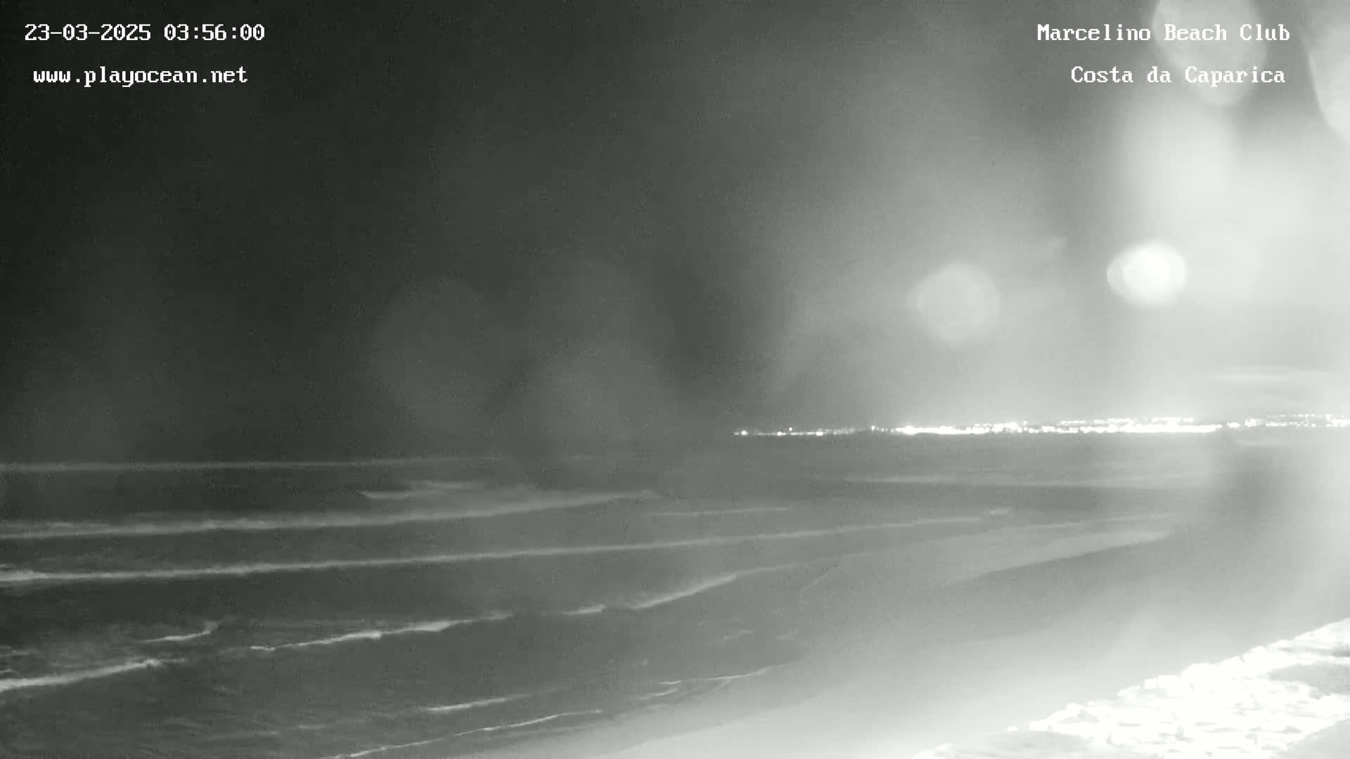 A nighttime view of a beach with gently rolling waves under a dark sky, showing a distant shoreline with lights.
