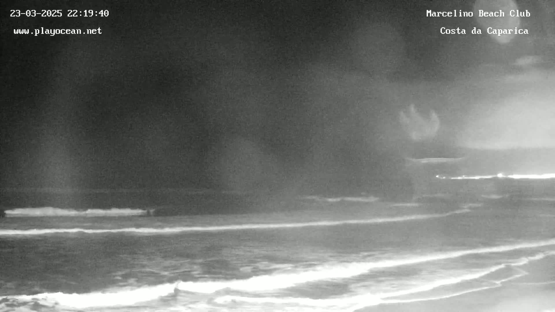 A nighttime, grayscale view of a dark ocean with small waves breaking on the shore under a cloudy sky.