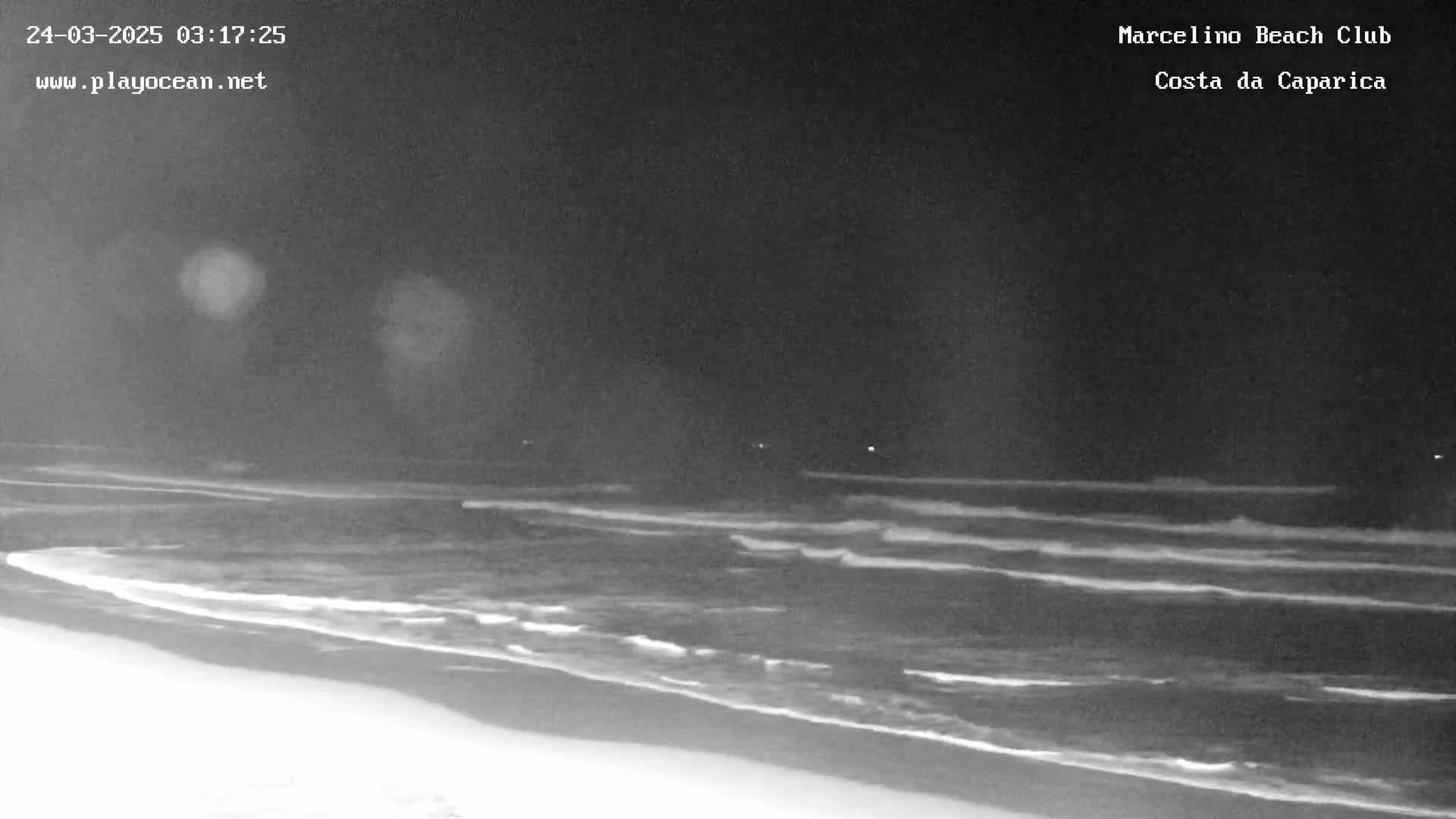 A dark, nighttime beach scene shows gentle waves lapping the shore under a clear sky.