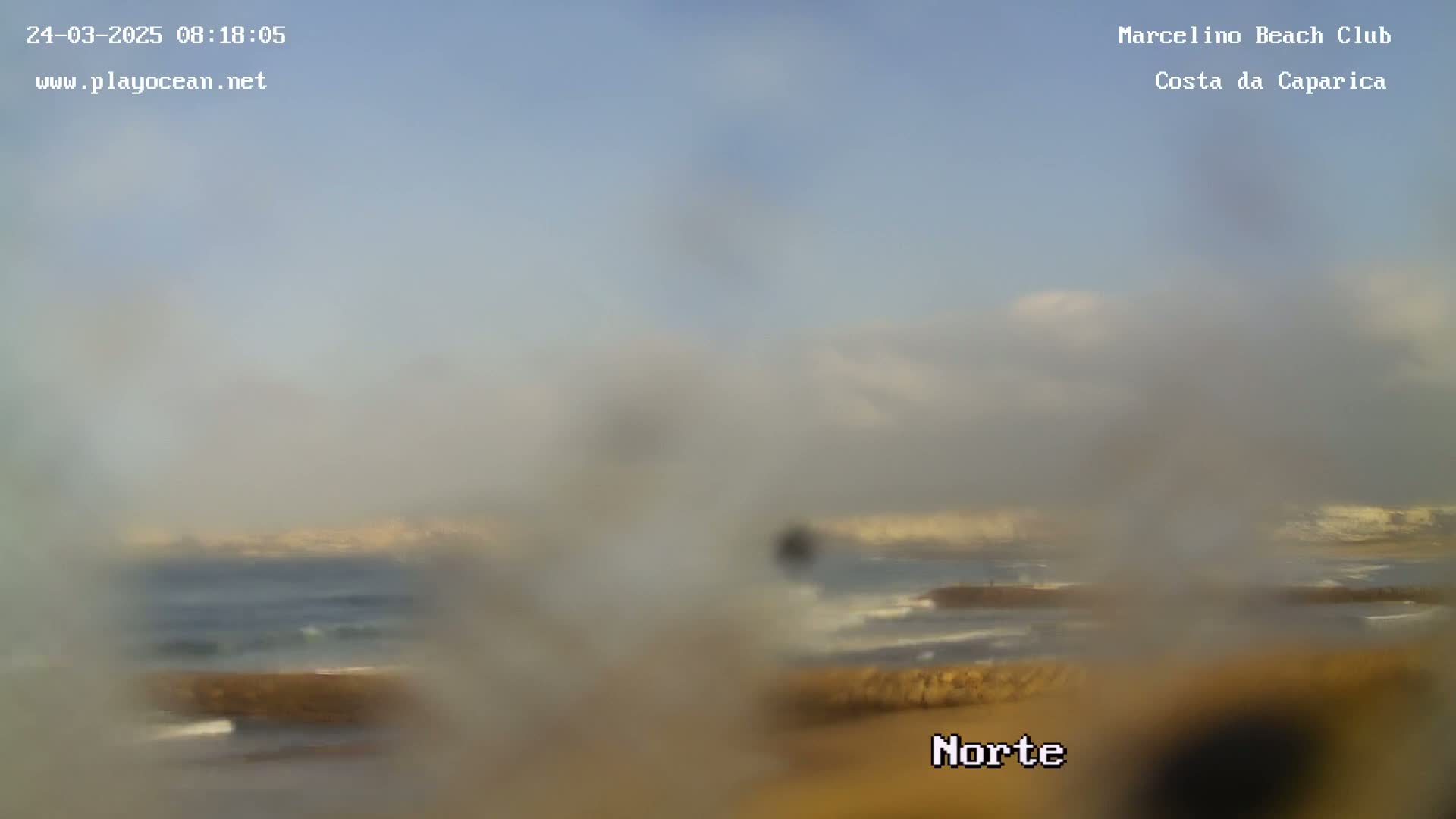 A hazy view of an ocean beach with gentle waves under a mostly cloudy sky.