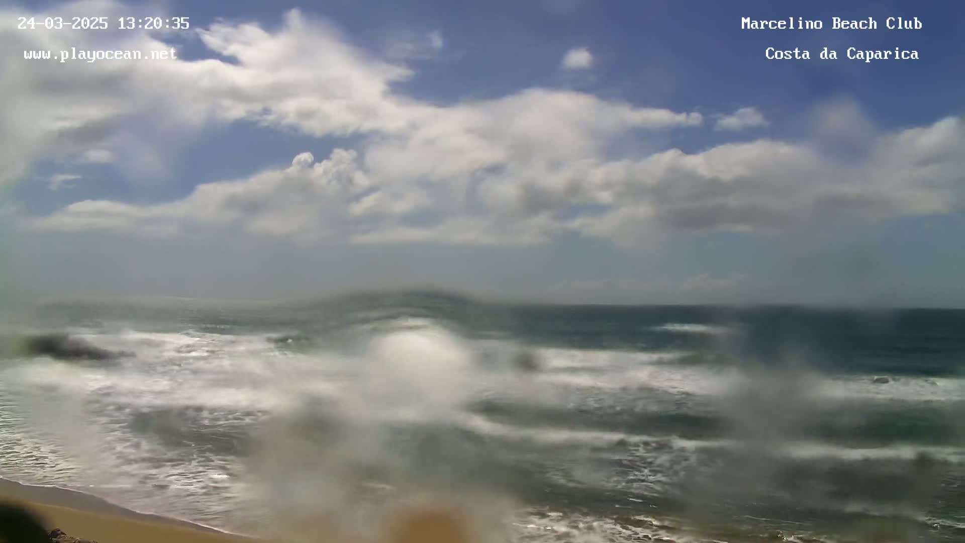 A partly cloudy day shows a somewhat blurry view of a beach with moderately sized waves crashing on the shore.