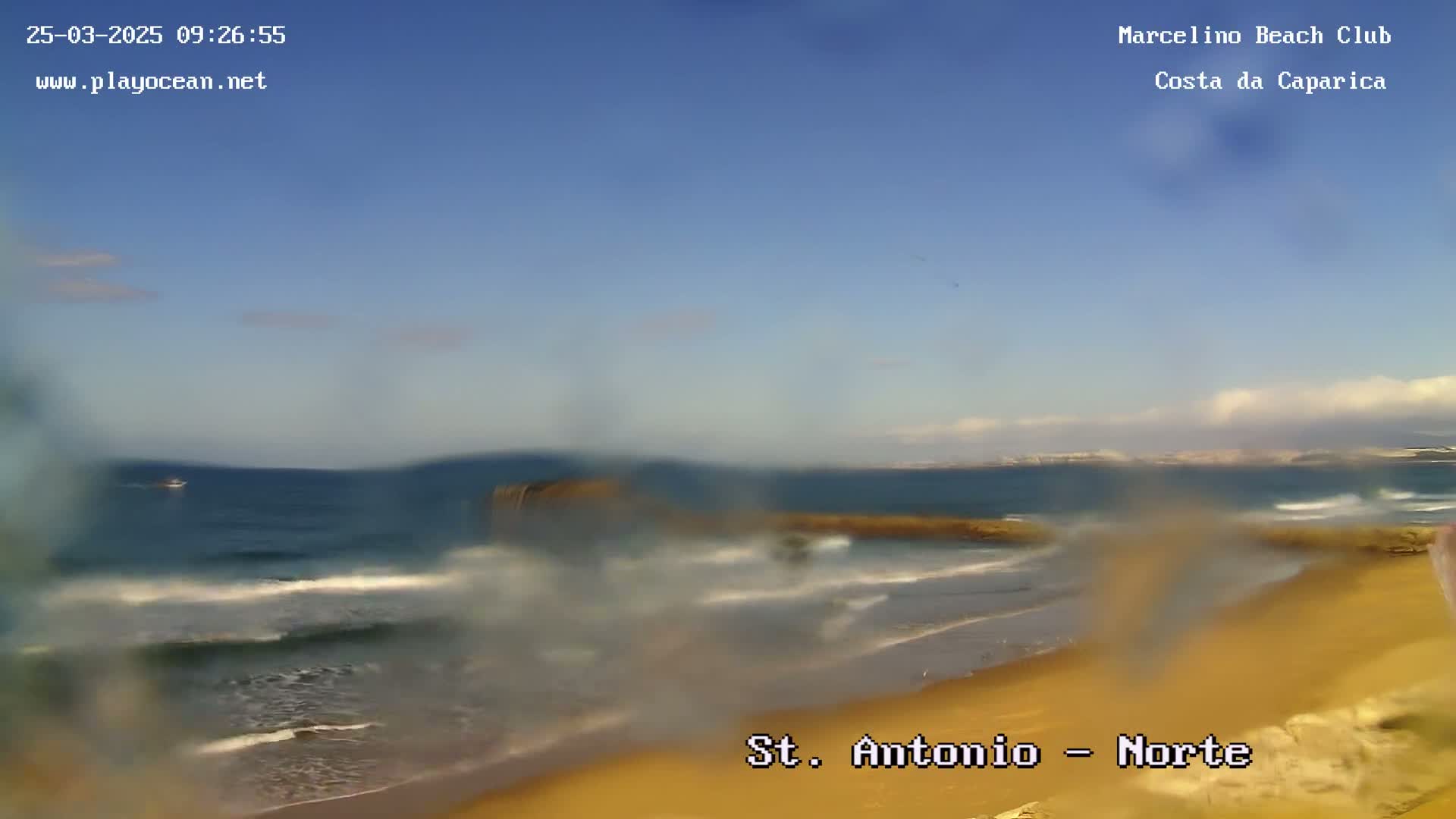 A hazy, sunny day at a beach shows a calm ocean with small waves lapping a sandy shore.