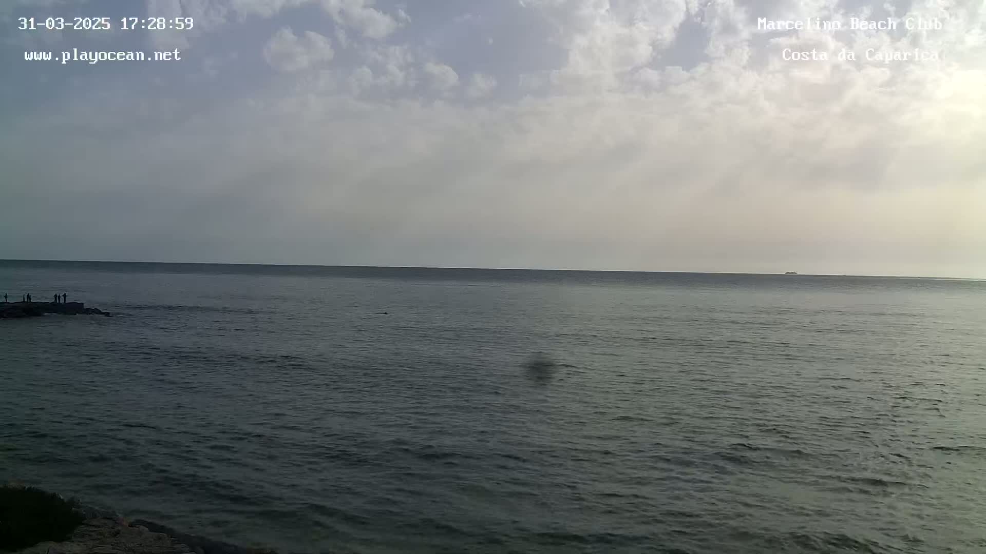 A mostly cloudy sky overlooks a calm ocean where a few people stand on rocks near the shore.