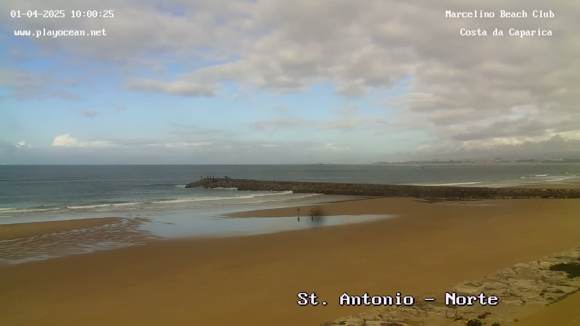 A sandy beach with a long, low rock barrier extending into the ocean under a partly cloudy sky.