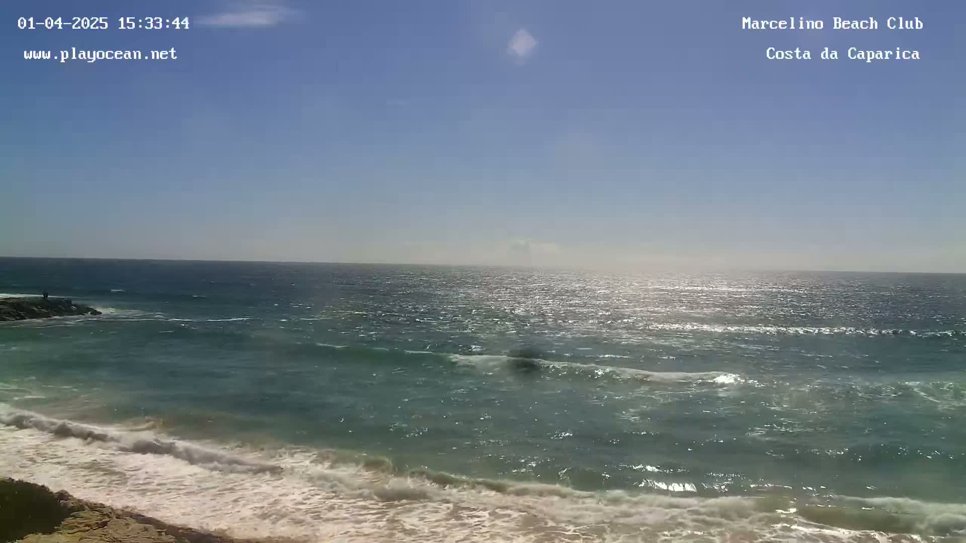 A sunny day at a beach shows a calm ocean with small waves breaking on the shore.