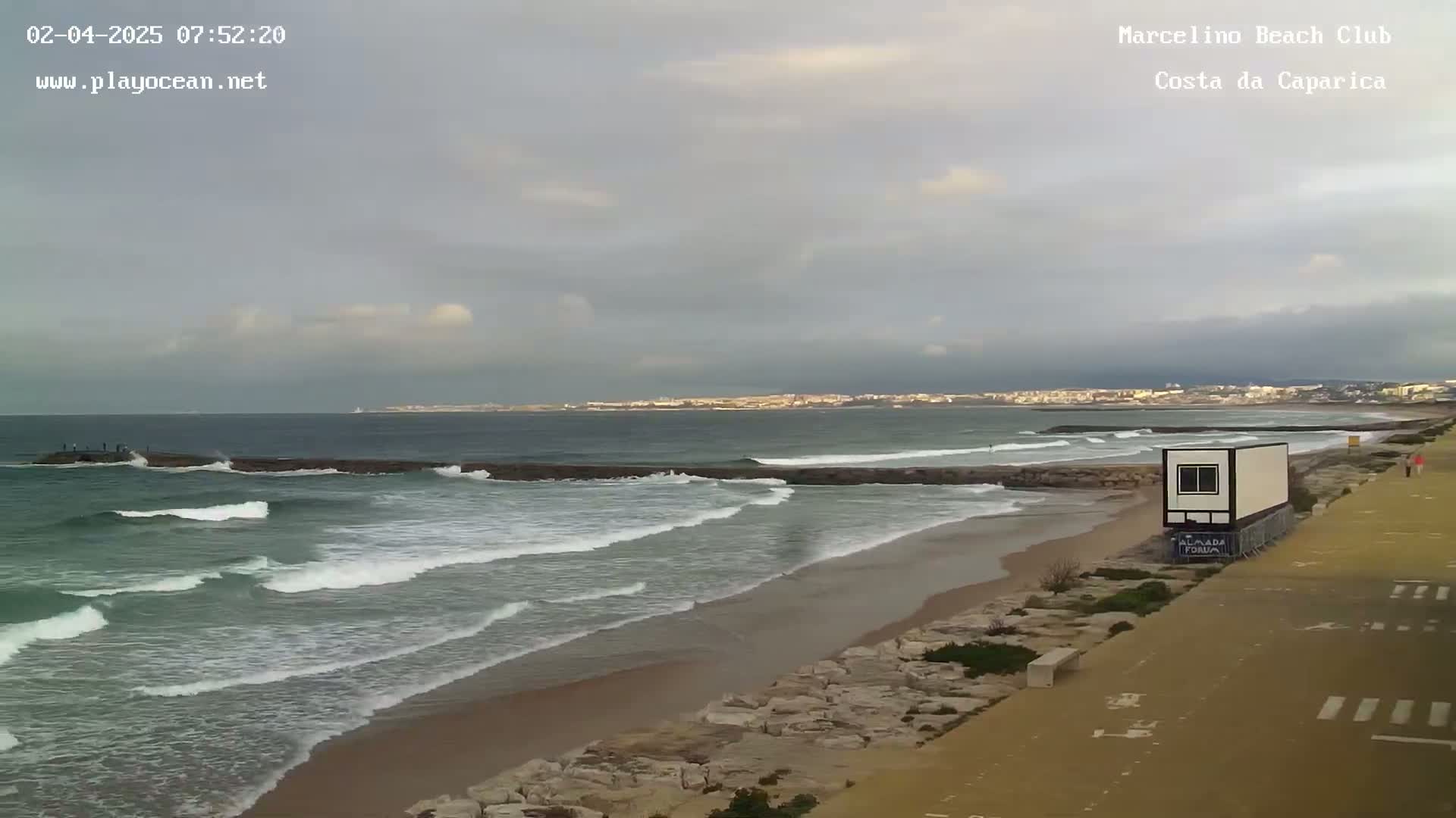 A sandy beach with moderate waves under an overcast sky shows a distant town in the background.