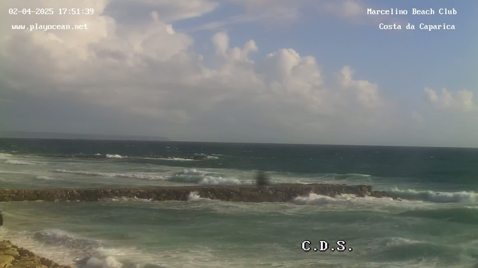 A partly cloudy day shows a rocky breakwater partially submerged by choppy ocean waves.