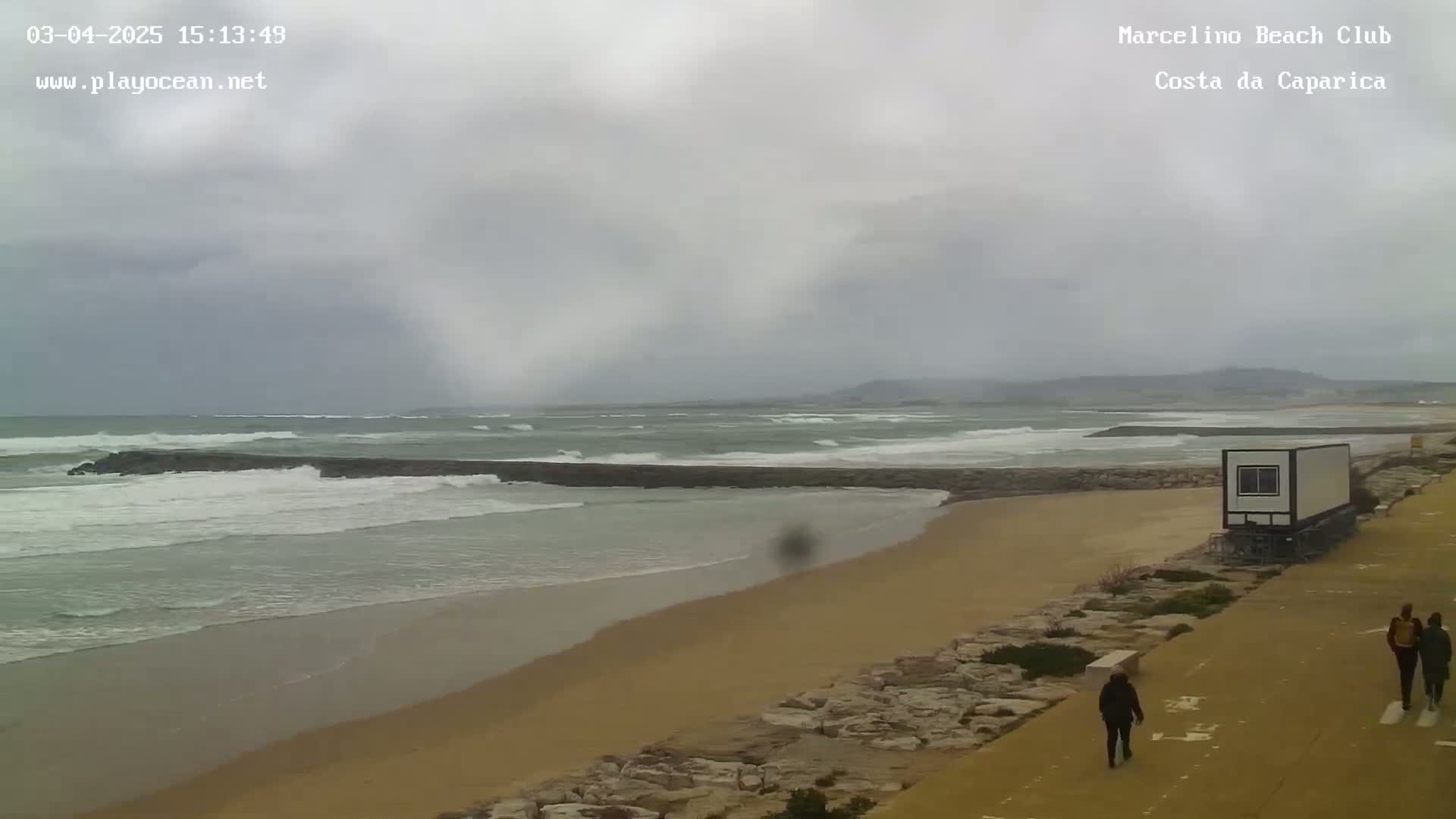 A sandy beach with choppy waves under an overcast sky shows a few people walking along a paved path next to a small, white building.