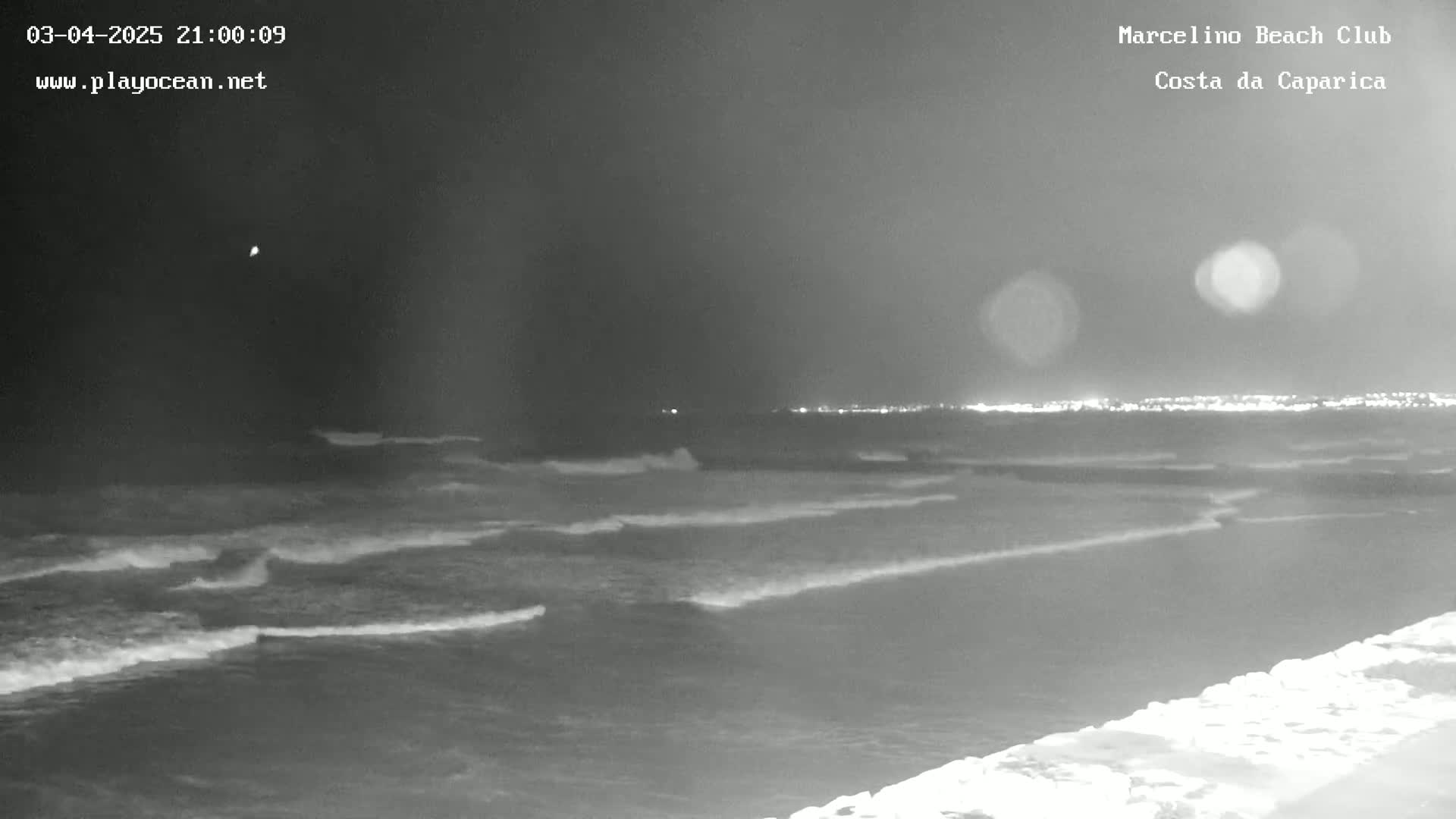 A nighttime, grayscale image shows moderate waves lapping at a shore under a dark sky.