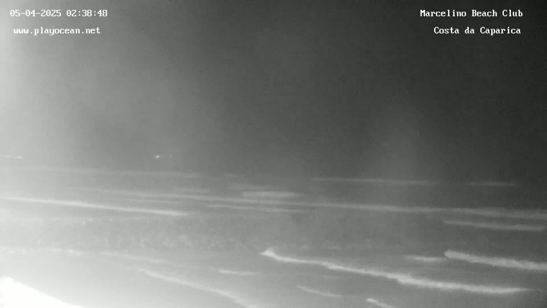 A nighttime, low-light view shows a dark beach with gently rolling waves under a cloudy sky.