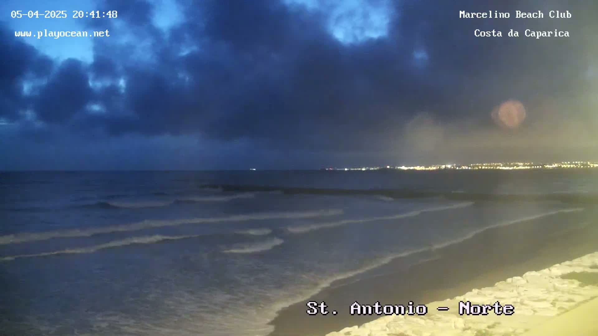 Gentle waves lap a dark beach under a mostly cloudy, twilight sky, with city lights visible in the distance.