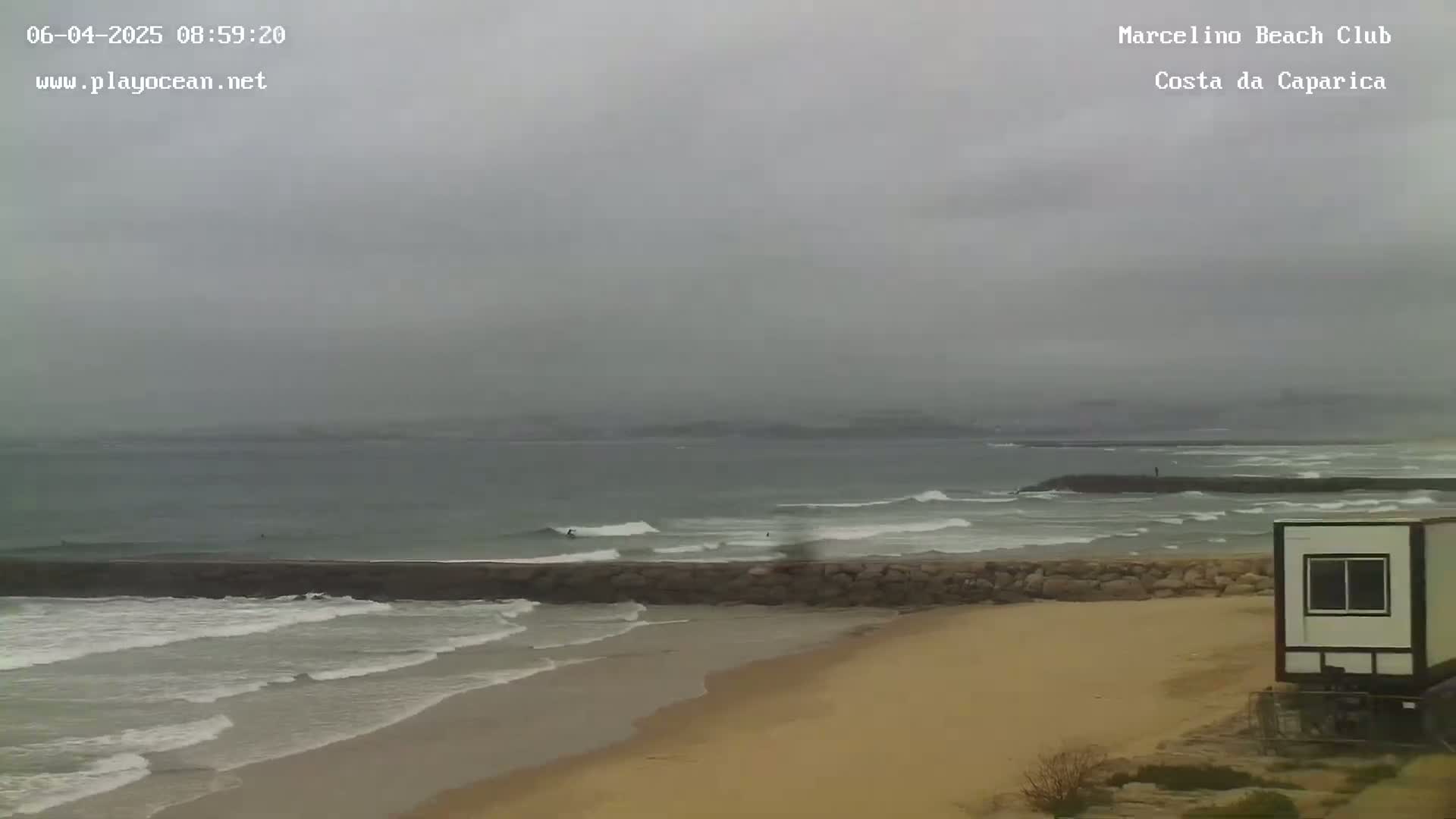 A sandy beach with a low stone wall separating it from a grey, overcast ocean with small waves and a few surfers.