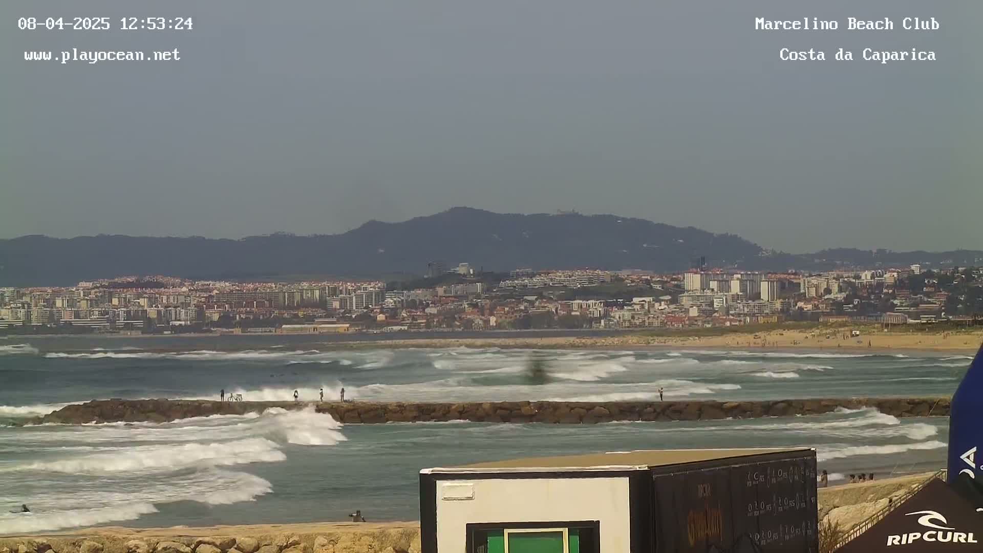 A coastal city is visible in the distance under a hazy sky, with moderate waves breaking against a rocky jetty in the foreground.
