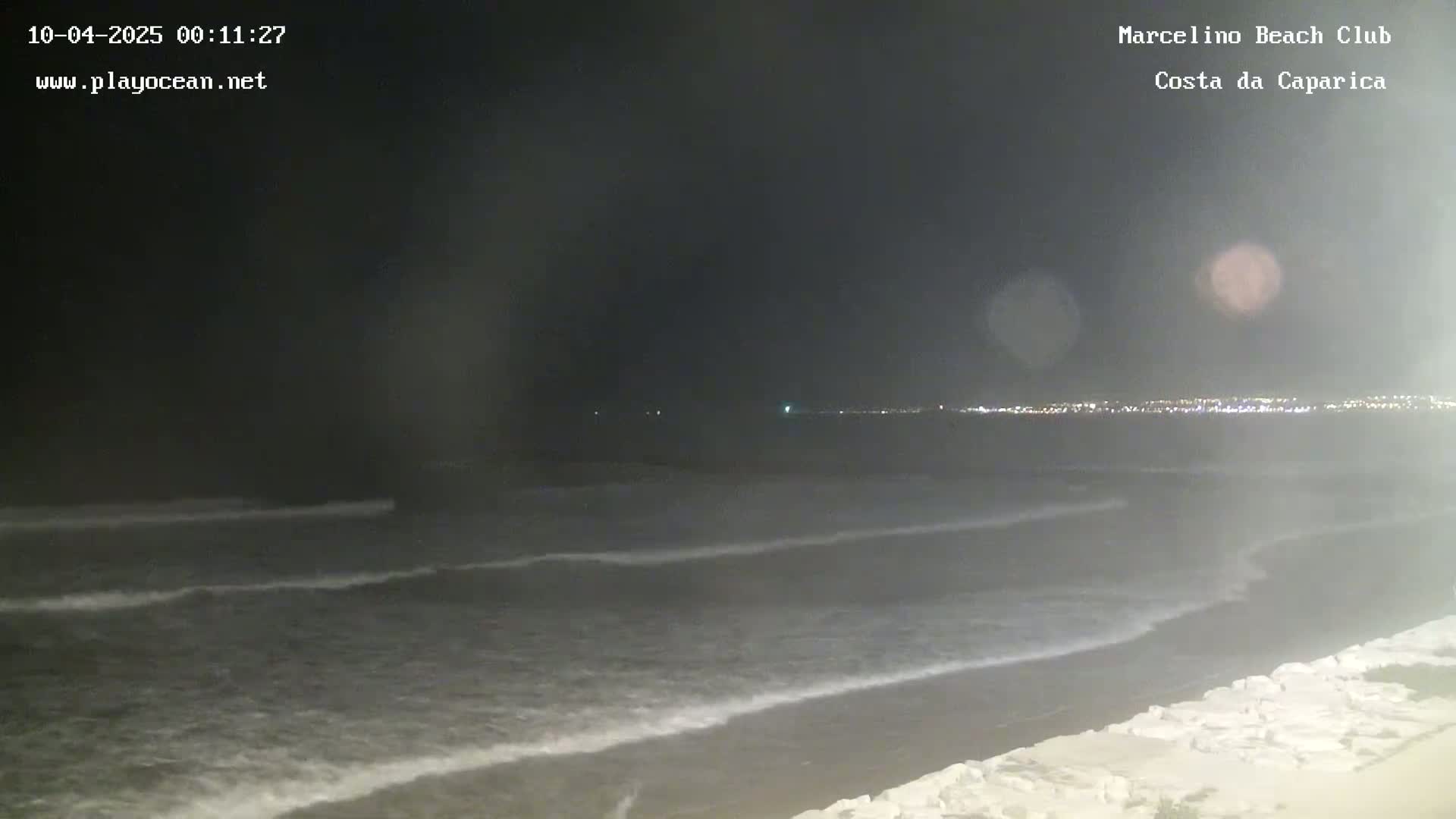 A dark, nighttime view of a beach with gently rolling waves and a city skyline faintly visible in the distance.
