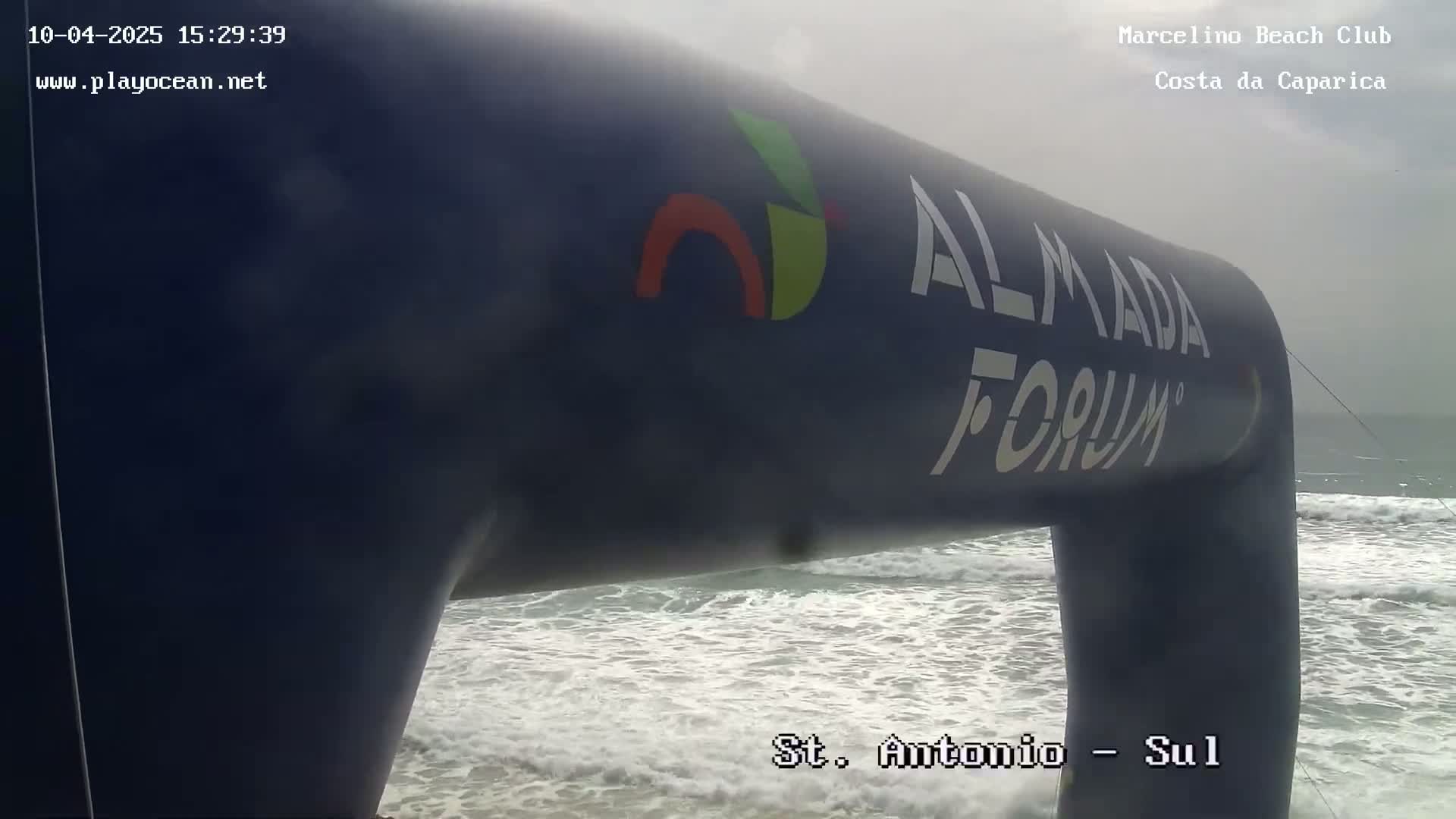 A large, dark-blue inflatable arch stands on a beach with moderate waves under an overcast sky.
