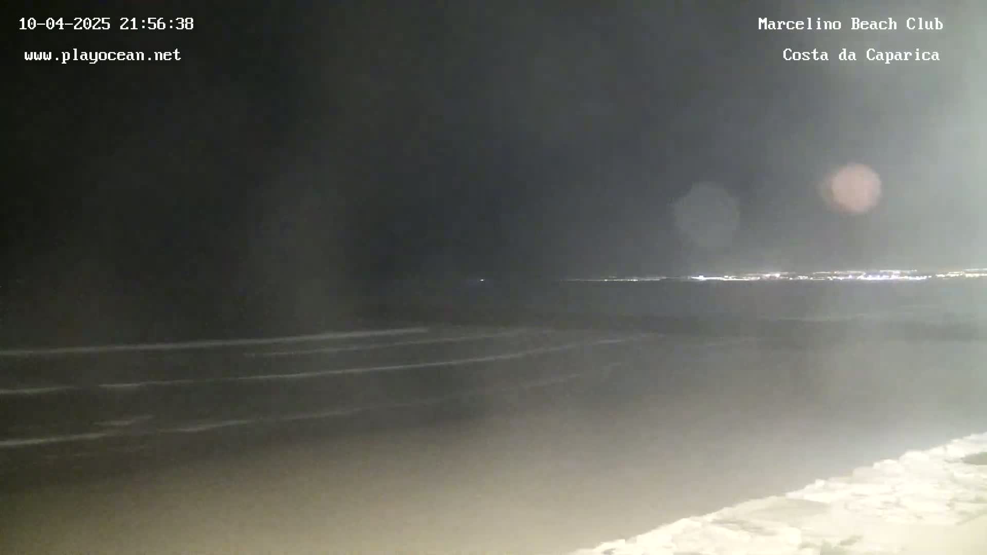 A dark, nighttime view of a beach with gently lapping waves and a distant city skyline faintly illuminated.