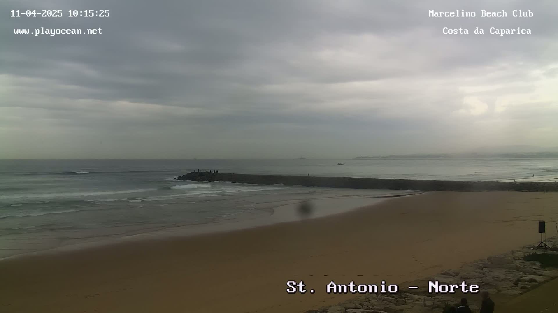 A sandy beach with a rock jetty extends into a gray, overcast ocean with small waves.