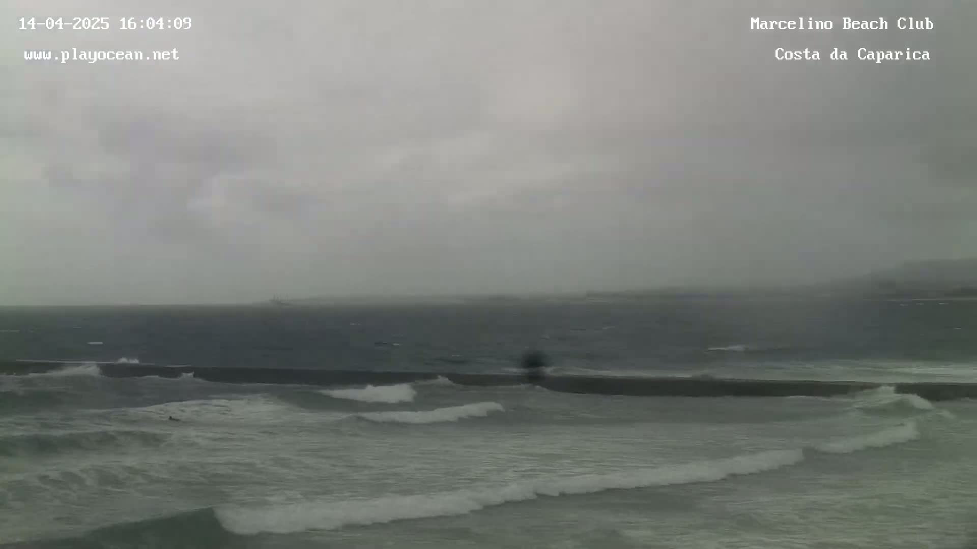 A dark, overcast sky hangs over a choppy ocean with waves breaking against a low wall.