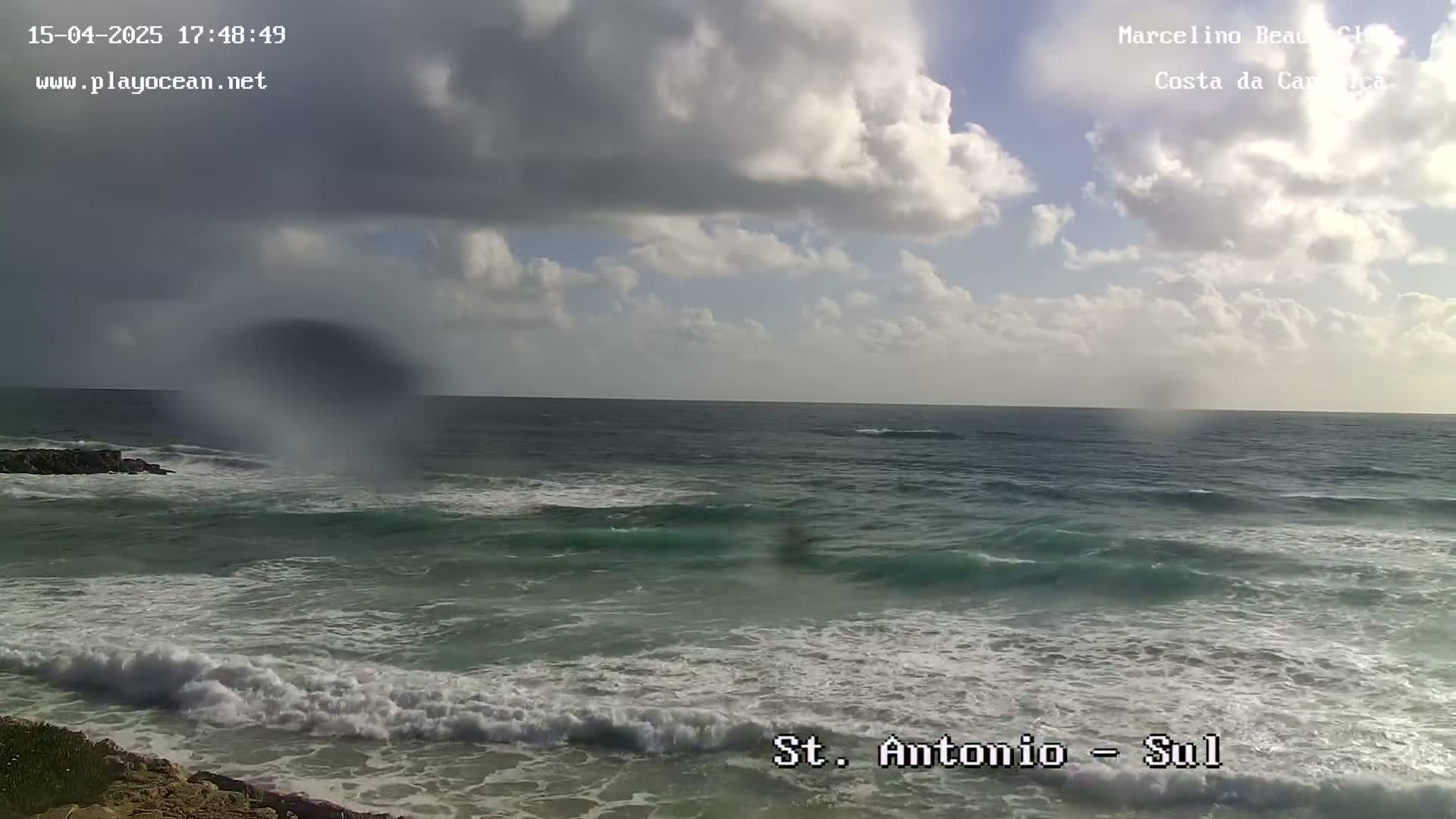 A partly cloudy day shows moderate waves crashing on a sandy shore, with some rain obscuring the view.
