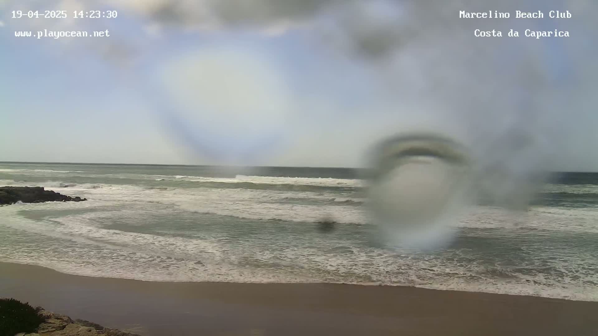 A view of a beach with moderate waves under an overcast sky, seen through a rain-covered lens.