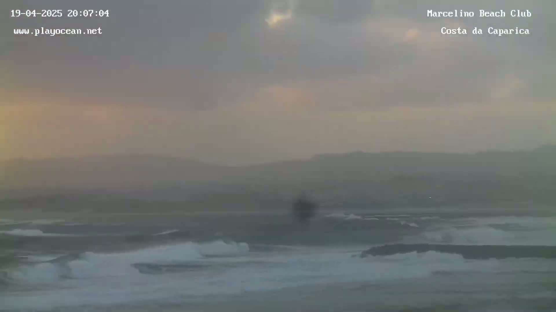 A hazy, overcast sky hangs over a beach with moderate waves rolling onto the shore.