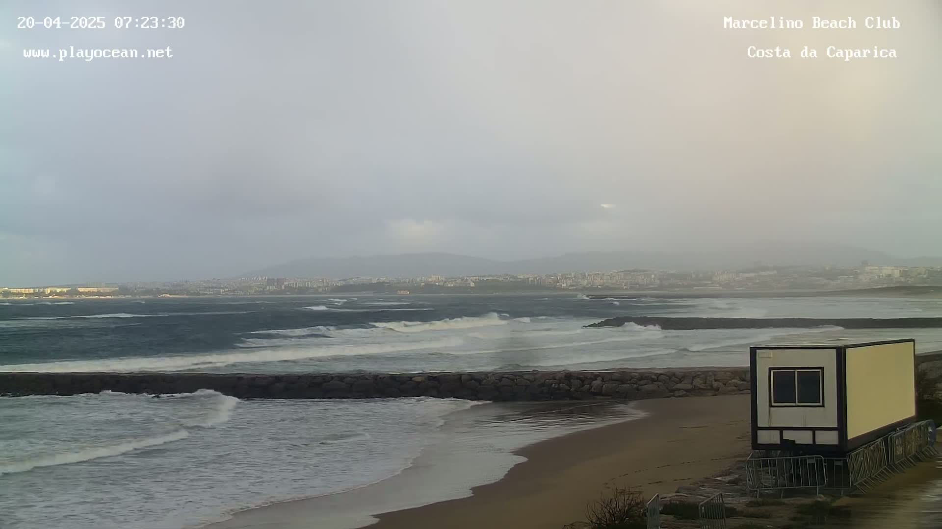 A sandy beach with choppy waves under an overcast sky, a distant city, and a small building near the shore.