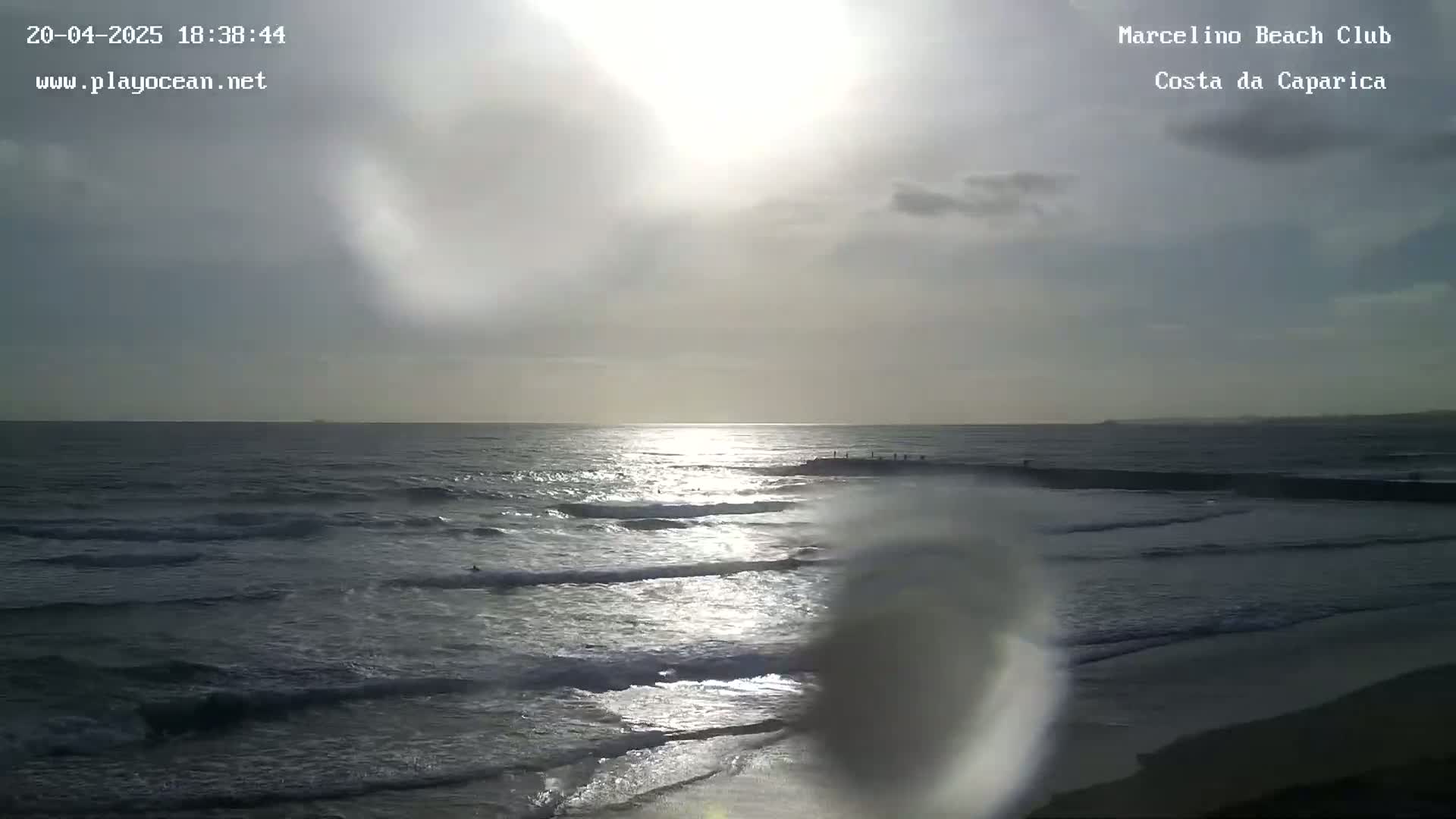 Under a partly cloudy sky, the sun shines on a wavy ocean with some people visible in the distance near a breakwater.