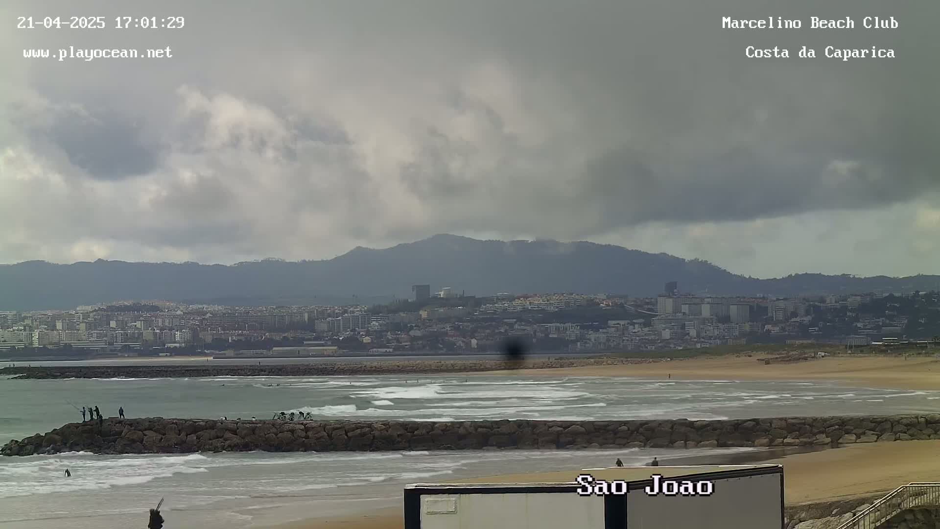 A cloudy day at a beach shows a calm ocean with small waves lapping against a rocky breakwater, several people fishing and surfing, and a distant cityscape nestled against mountains.