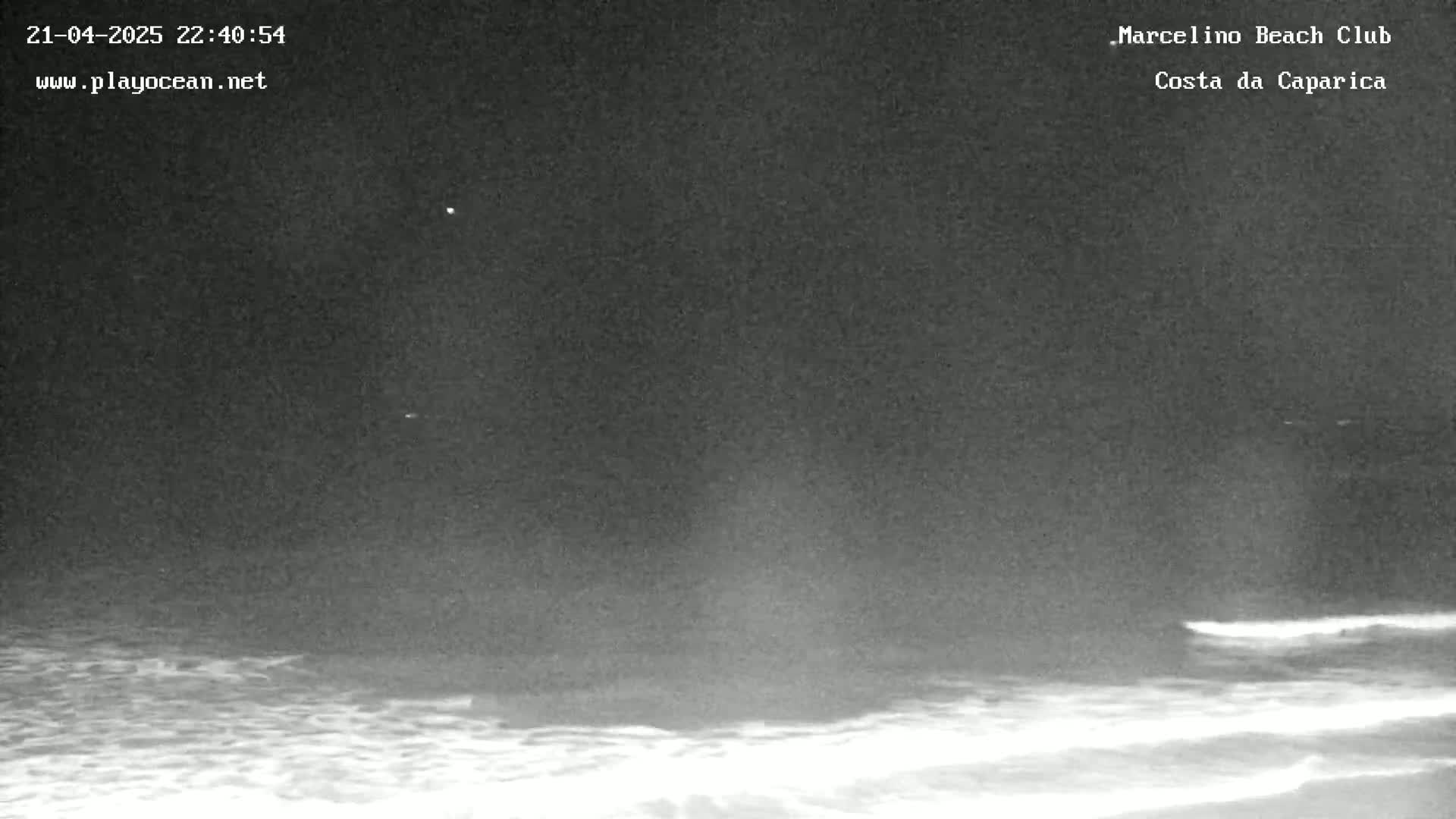 A nighttime, low-light view of a dark ocean with some waves breaking on the shore under a mostly clear sky.