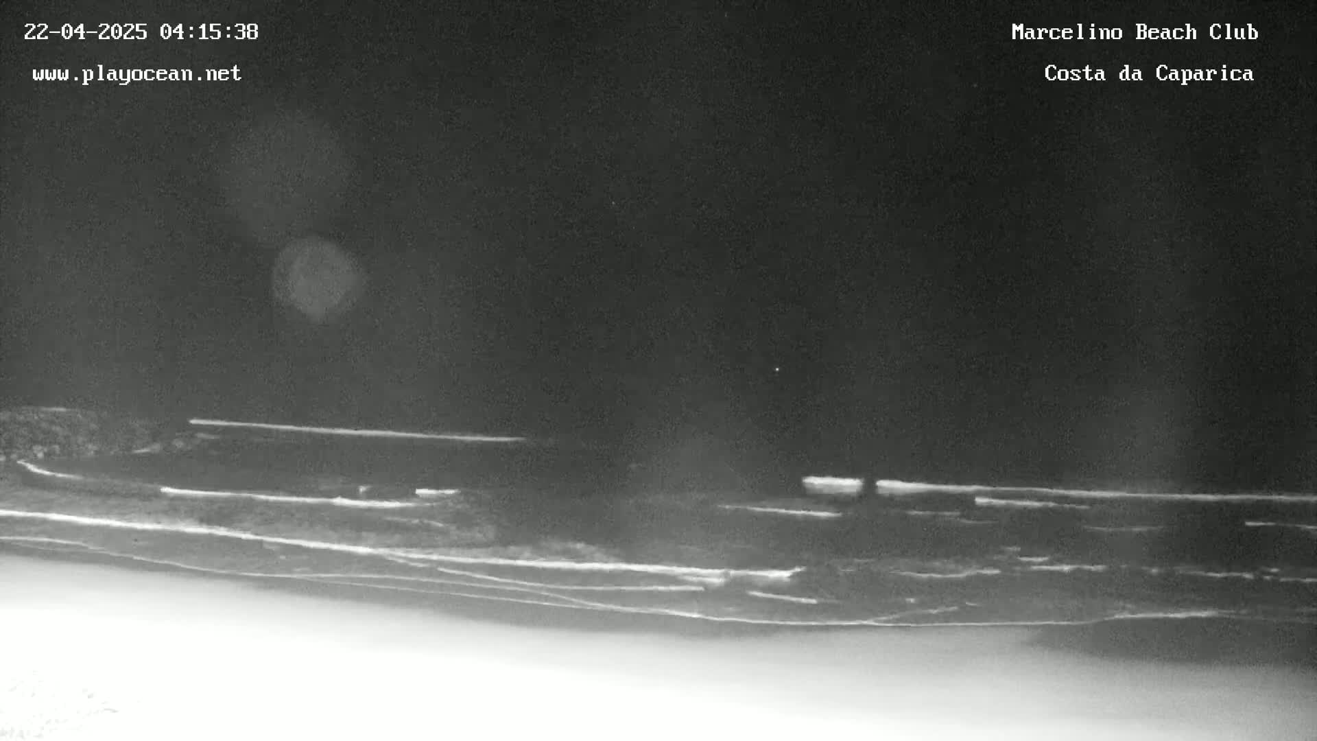 A nighttime, low-light image shows a dark beach and ocean with gently rolling waves under a clear, dark sky.