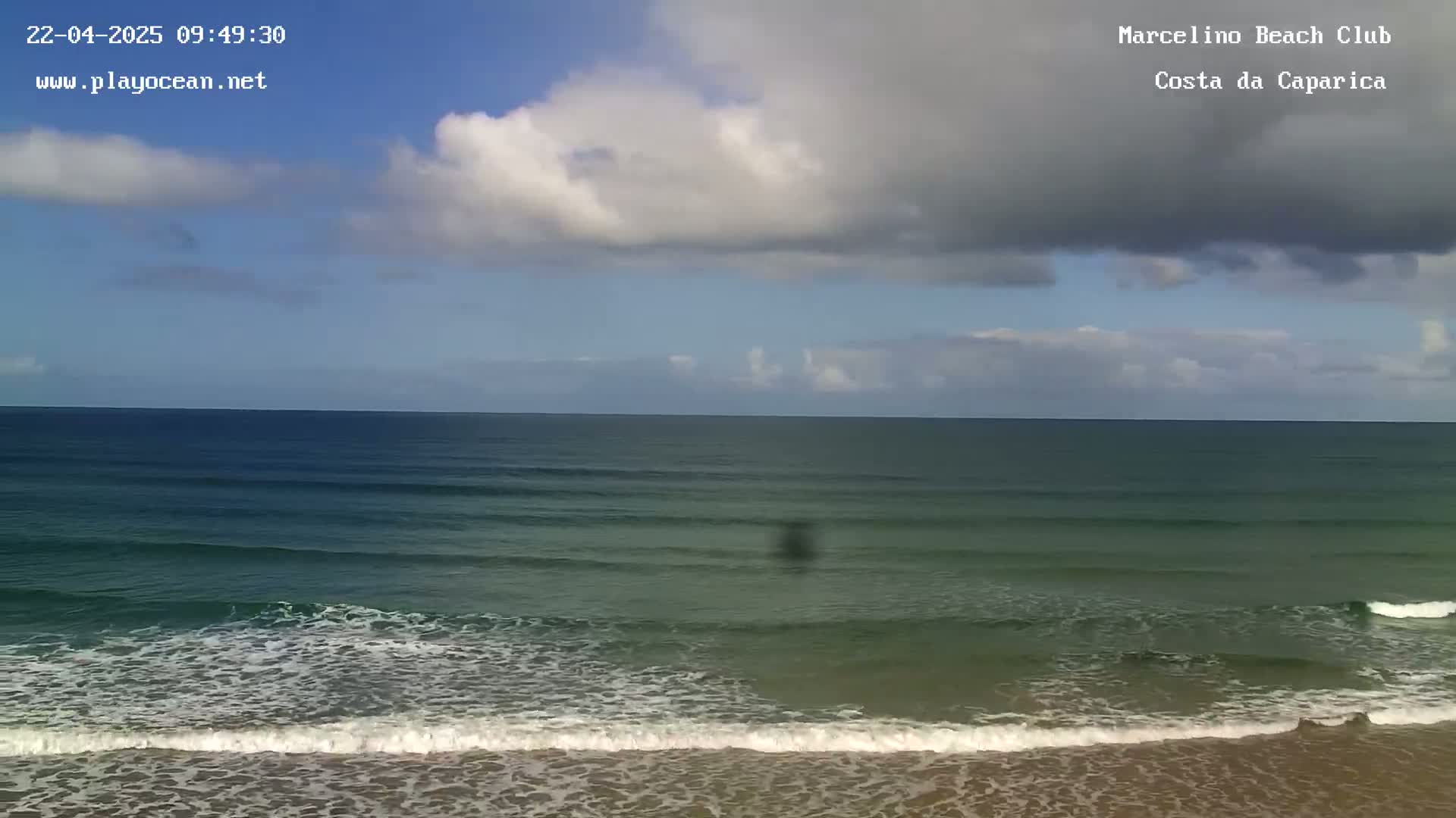 A partly cloudy sky overlooks a gently rolling ocean with small waves breaking on a sandy beach.