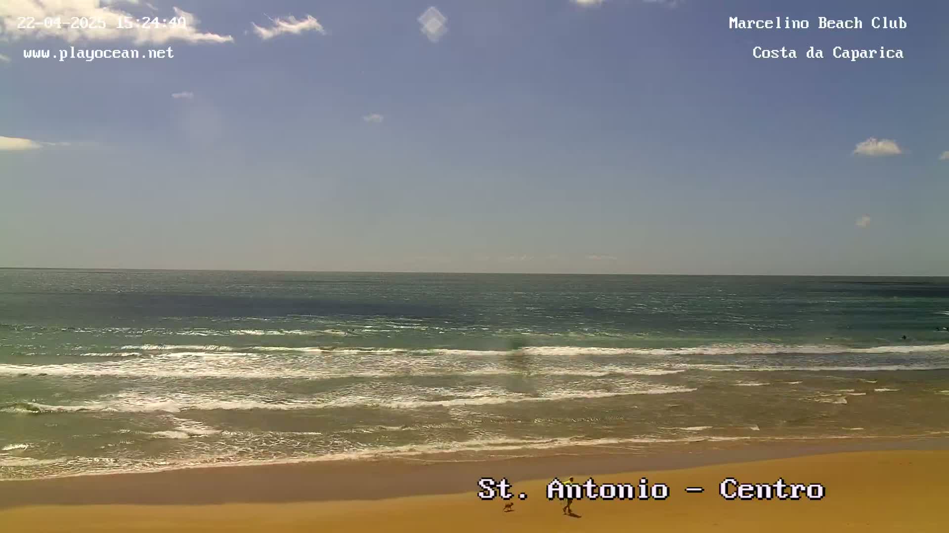 A sandy beach with gentle waves rolling onto the shore under a mostly clear, sunny sky.