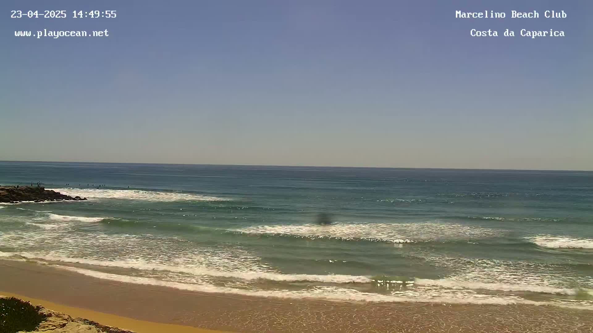 Under a clear blue sky, waves roll onto a sandy beach, with several people visible in the ocean.
