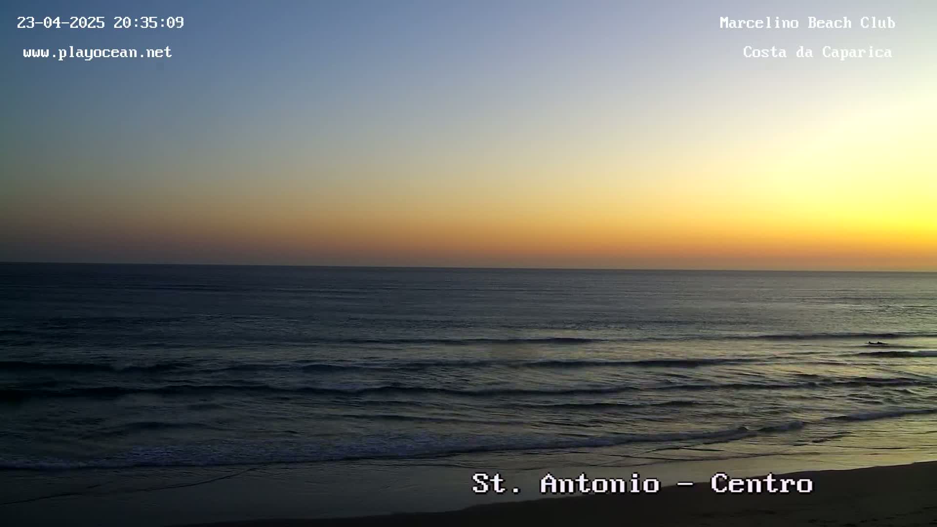 A dark ocean with small waves rolls onto a sandy beach under a sunset sky.