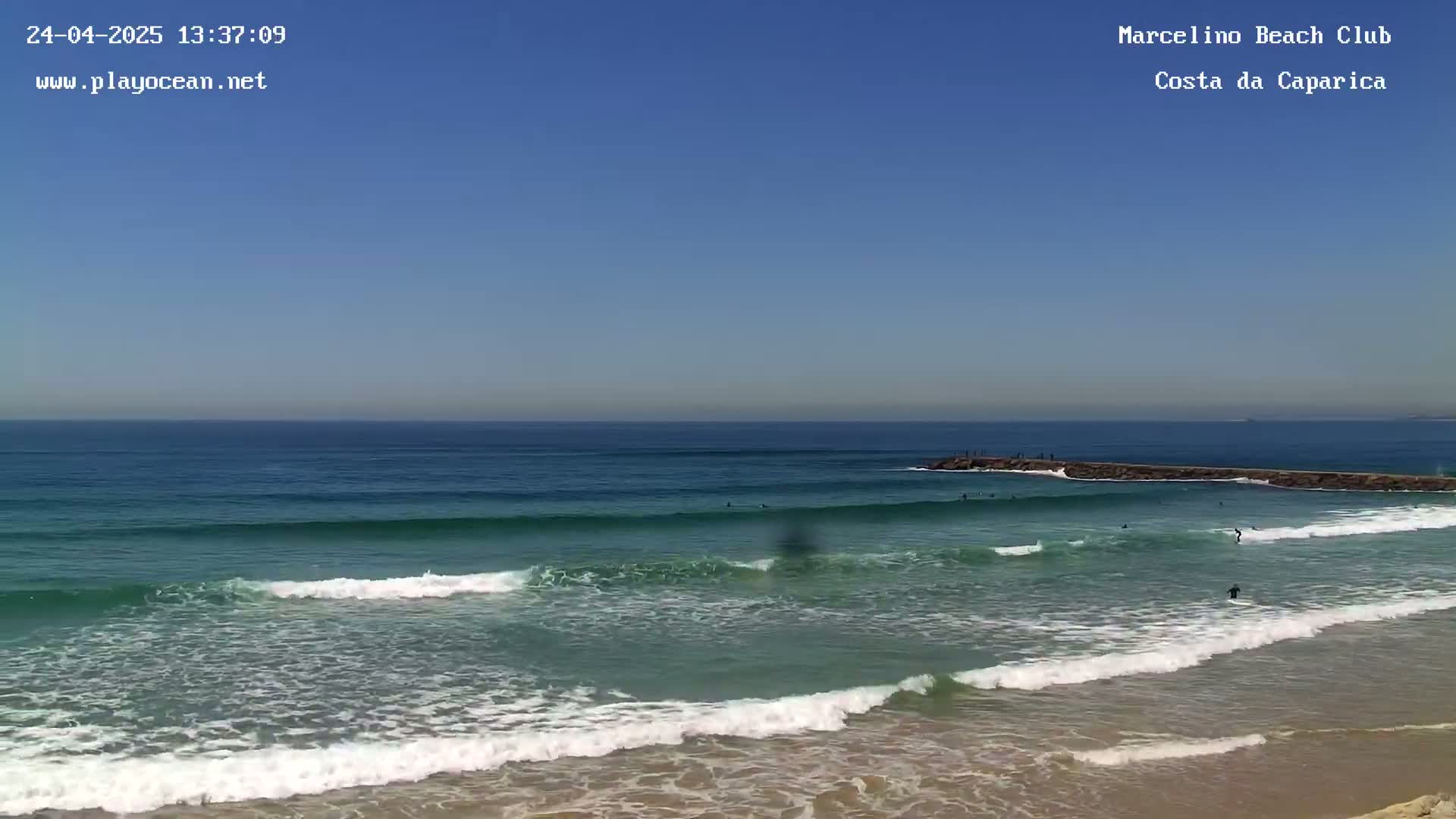 Under a clear blue sky, several surfers ride waves near a rock jetty at a sandy beach.