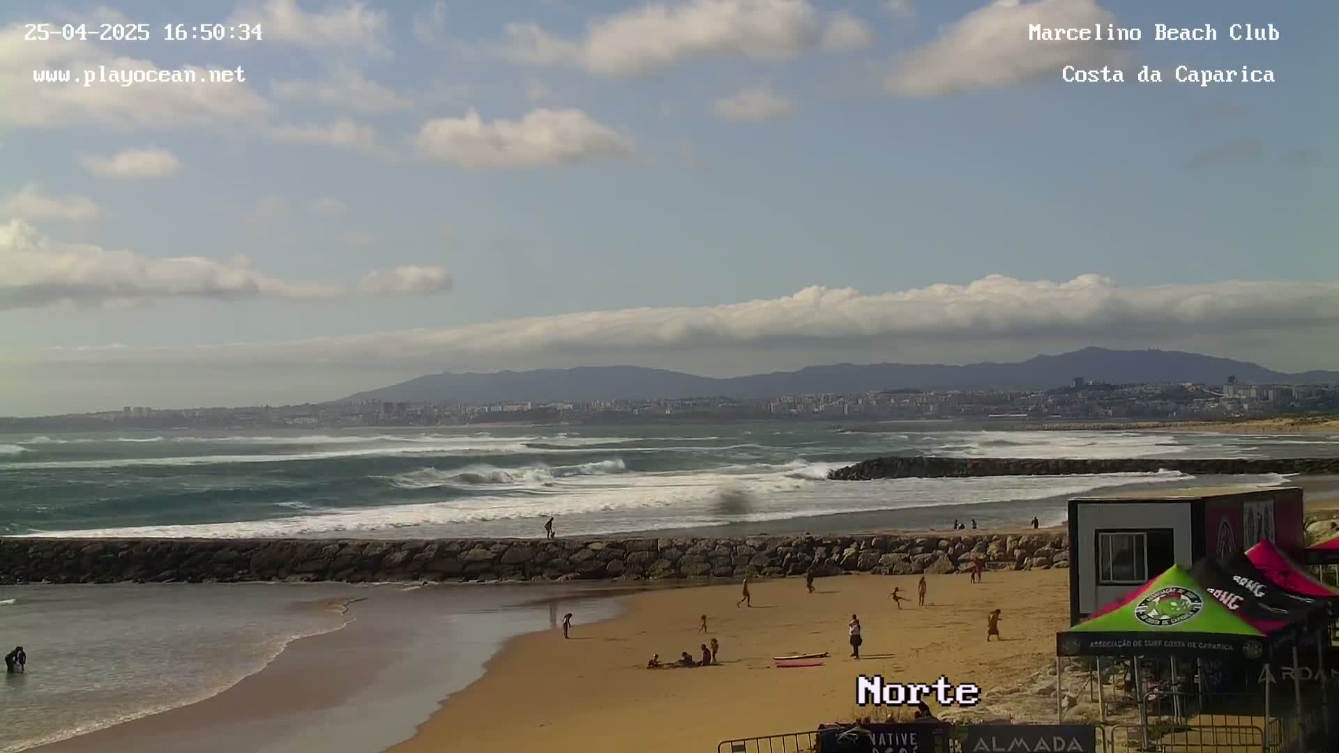 A partly cloudy day at a beach shows moderate waves breaking near a stone jetty, with people on the sandy shore.