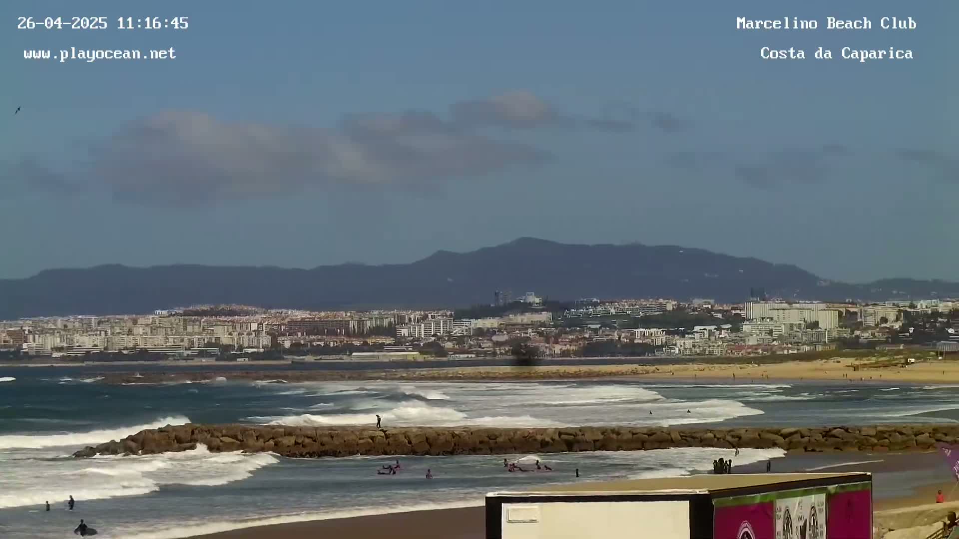 A partly cloudy day shows a beach with surfers in the water, a rocky breakwater, and a city in the background.