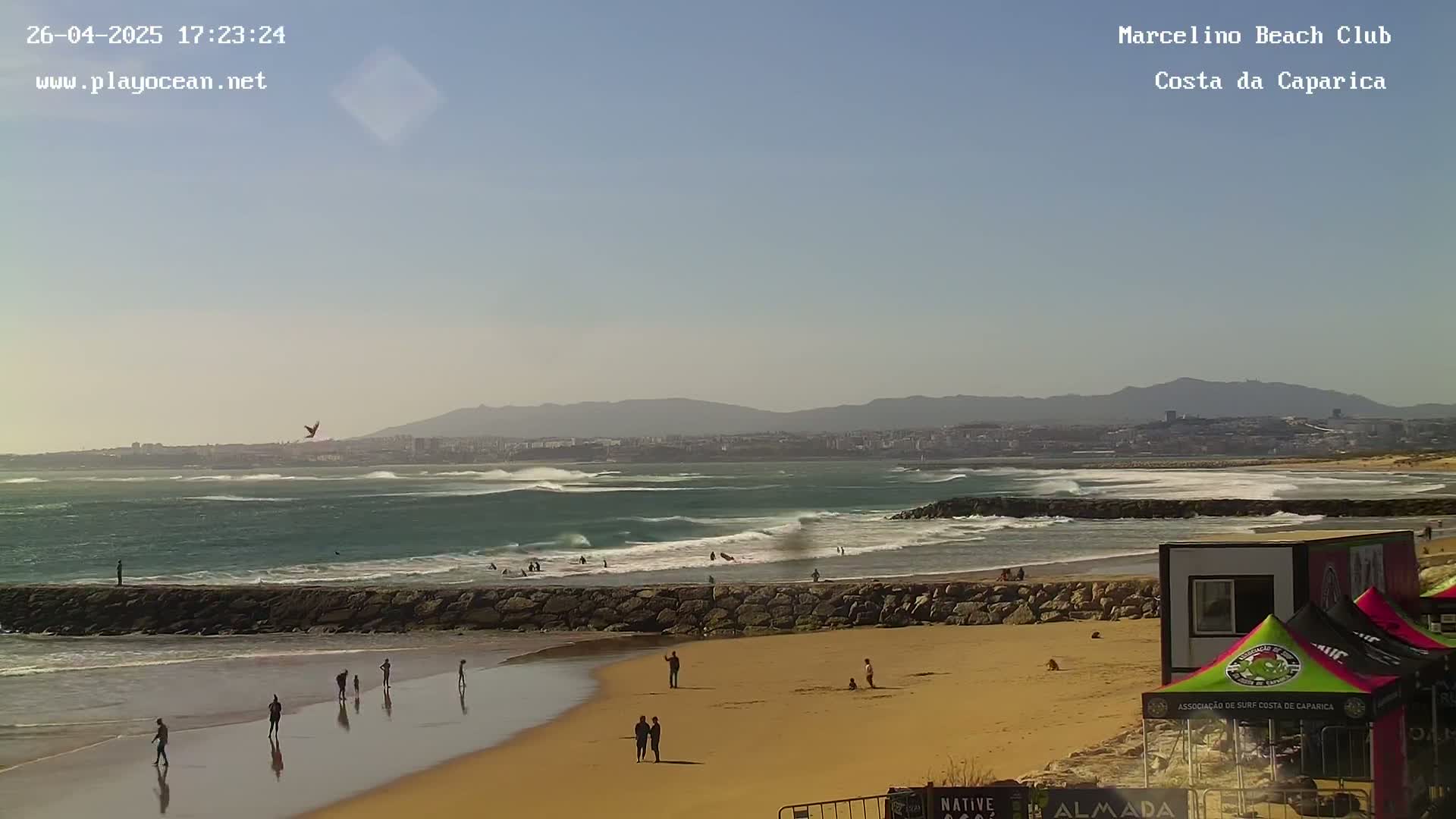 A sandy beach with a few people on it and some waves breaking in the ocean under a clear, sunny sky, with a distant city visible on the horizon.
