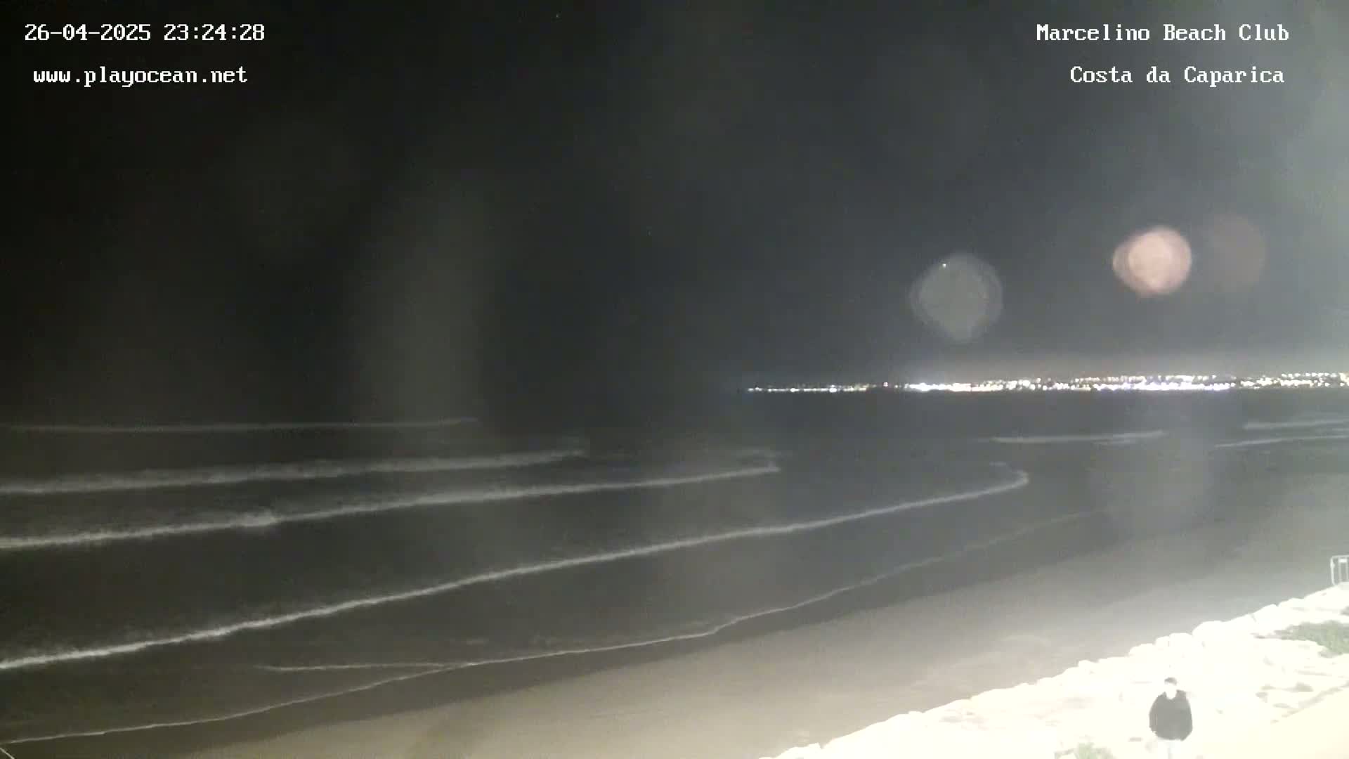A nighttime view of a dark ocean with small waves gently lapping the shore, under a clear, starlit sky, with city lights visible in the distance.
