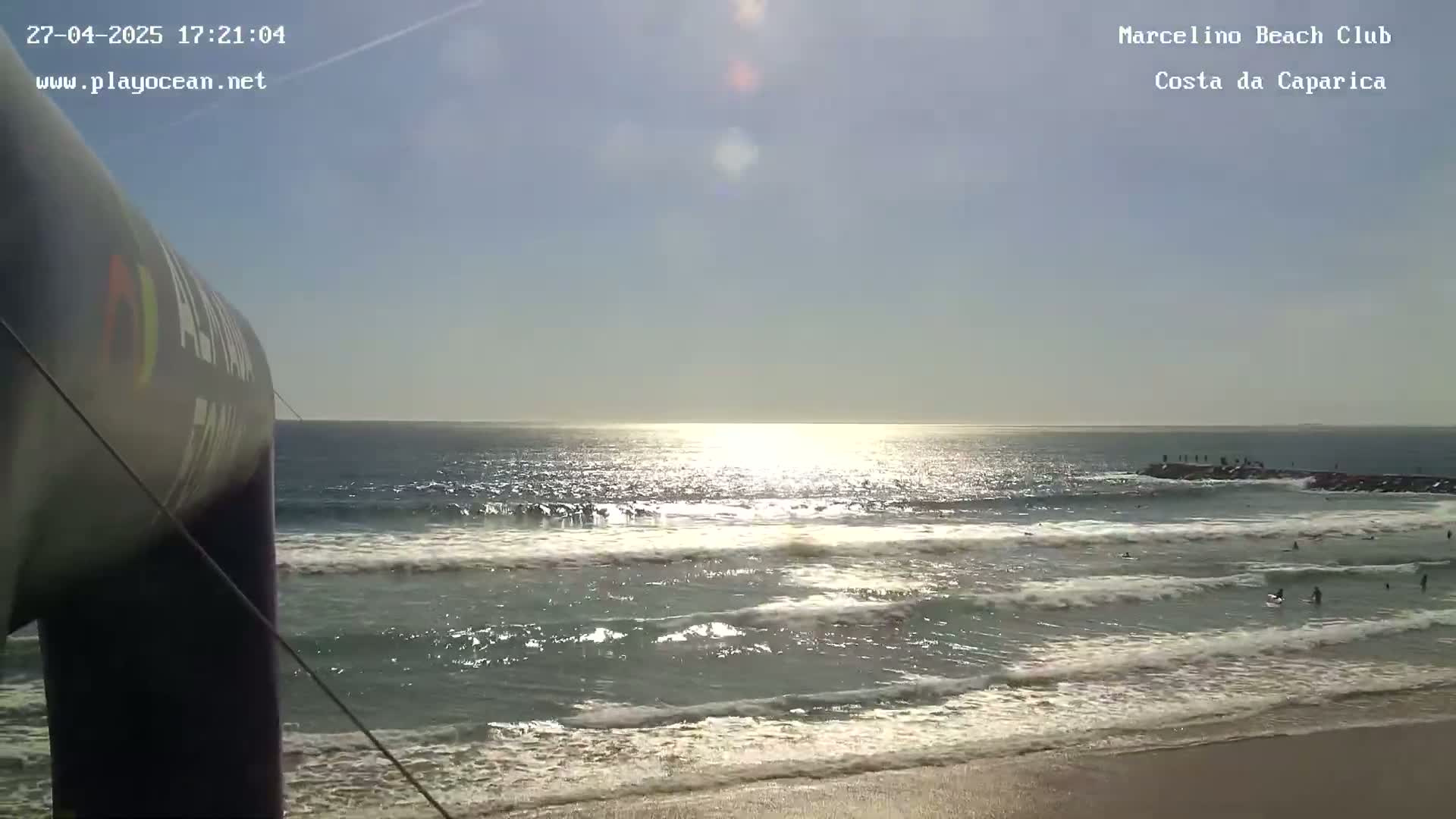 A sunny ocean beach scene shows waves rolling onto a sandy shore, with a few people in the water near a rock jetty.
