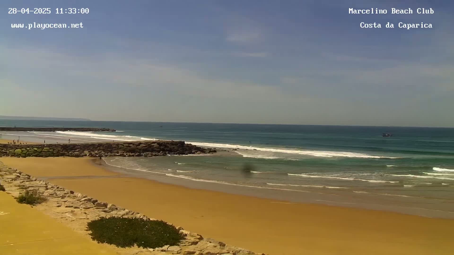 A sandy beach with small waves under a mostly clear sky stretches to a calm ocean.