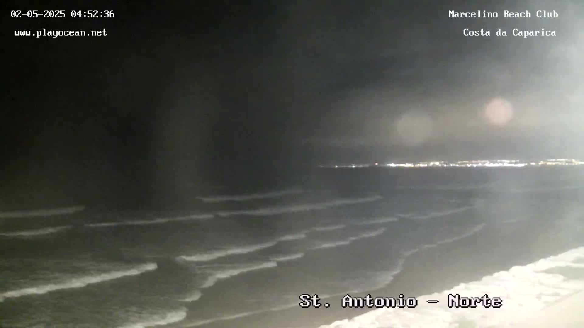 A nighttime view of a dark ocean with small waves rolling onto a sandy shore under a dark sky.